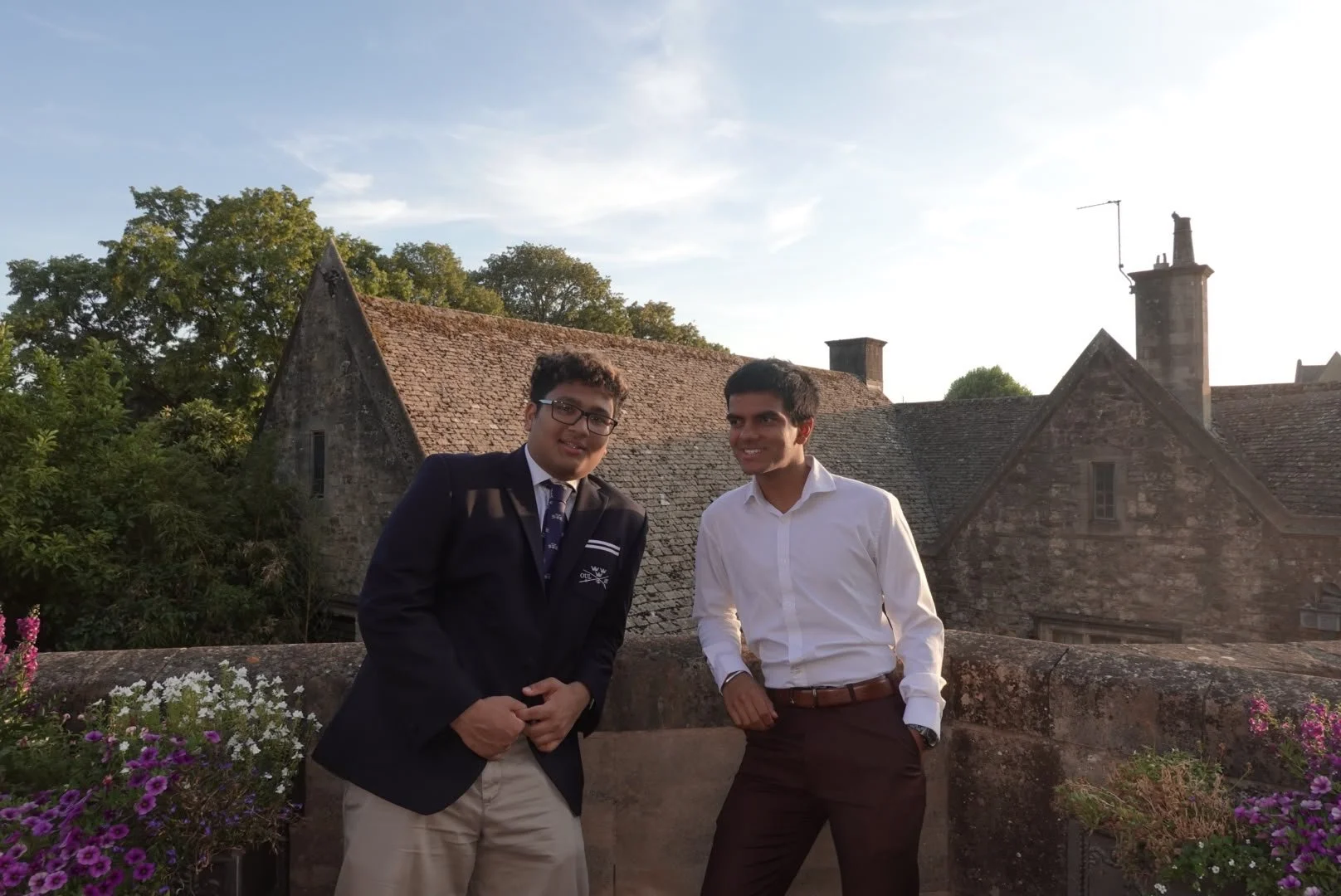 Two young men in formal attire standing on a stone balcony with floral arrangements, smiling, with historic stone buildings and trees in the background under a partly cloudy sky.