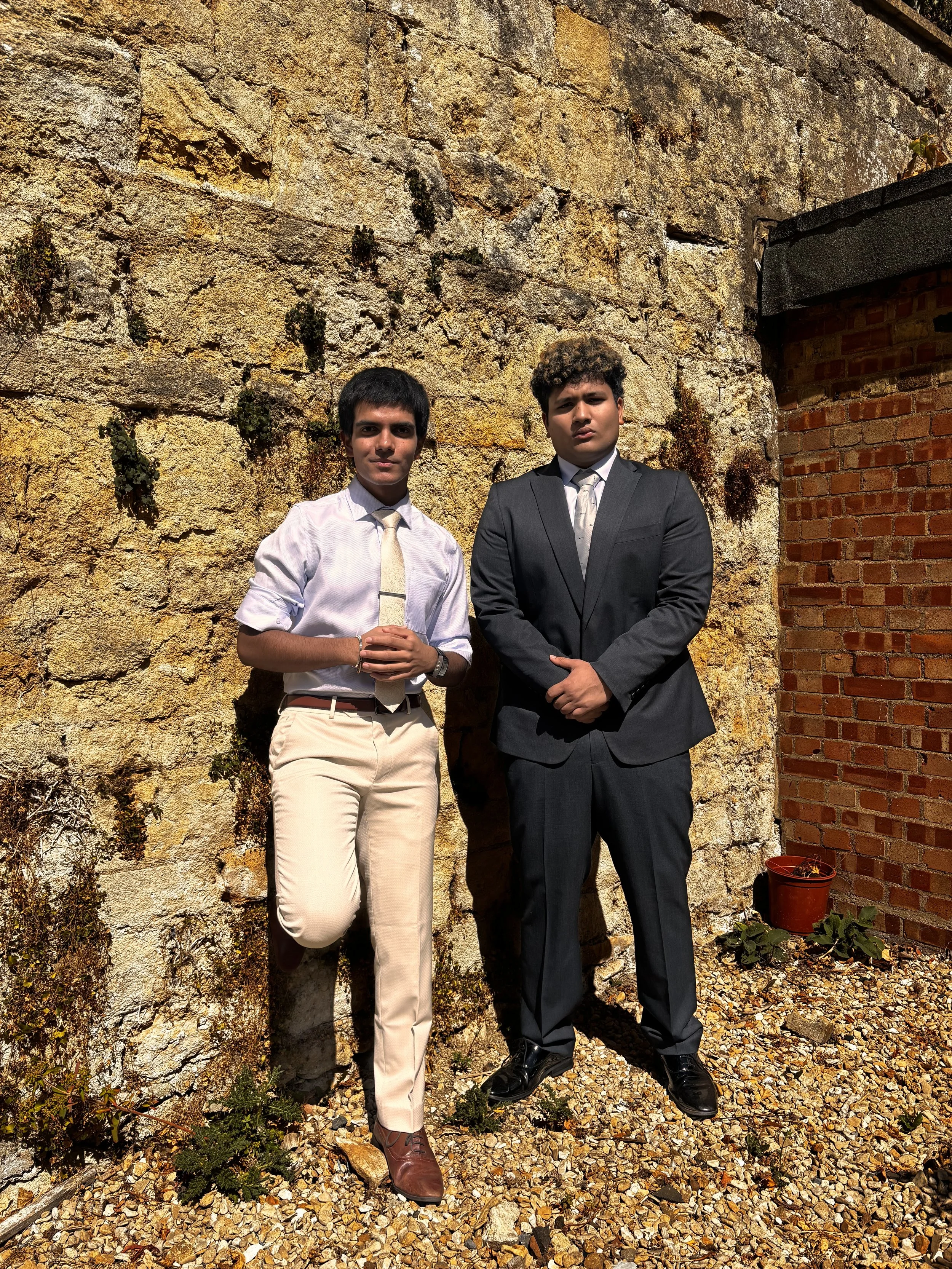 Two young men in formal attire standing outdoors against a textured stone wall with a small red flower pot on the ground nearby.