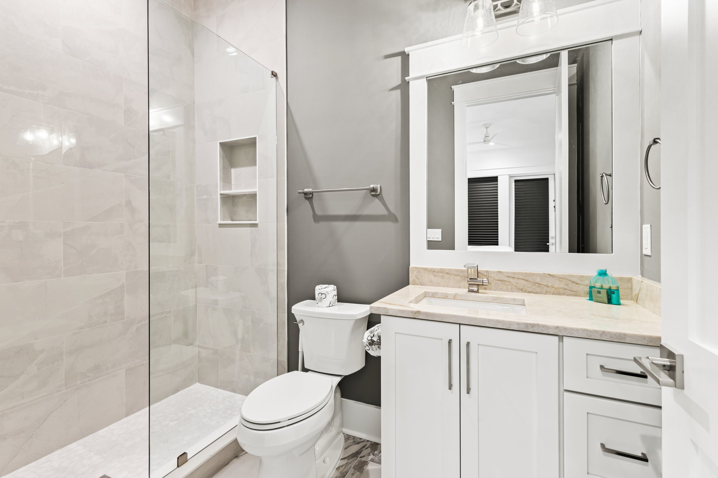 Modern bathroom with a frameless glass shower, toilet, and white vanity with a marble countertop, mirror, and above-counter sink.