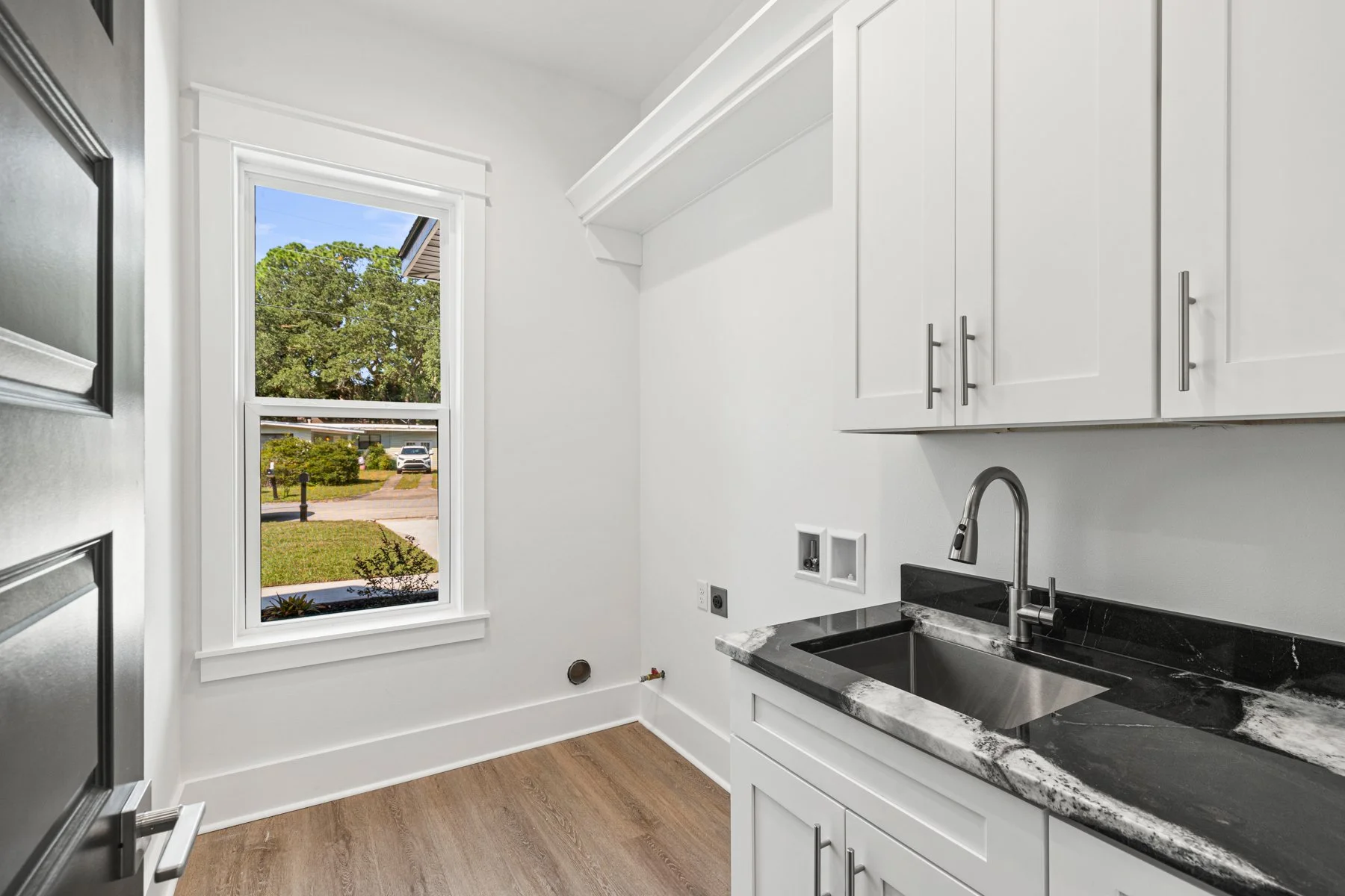 Modern white kitchen with a black marble countertop, sink, and white cabinets. Large window overlooking a suburban street with greenery and parked cars.