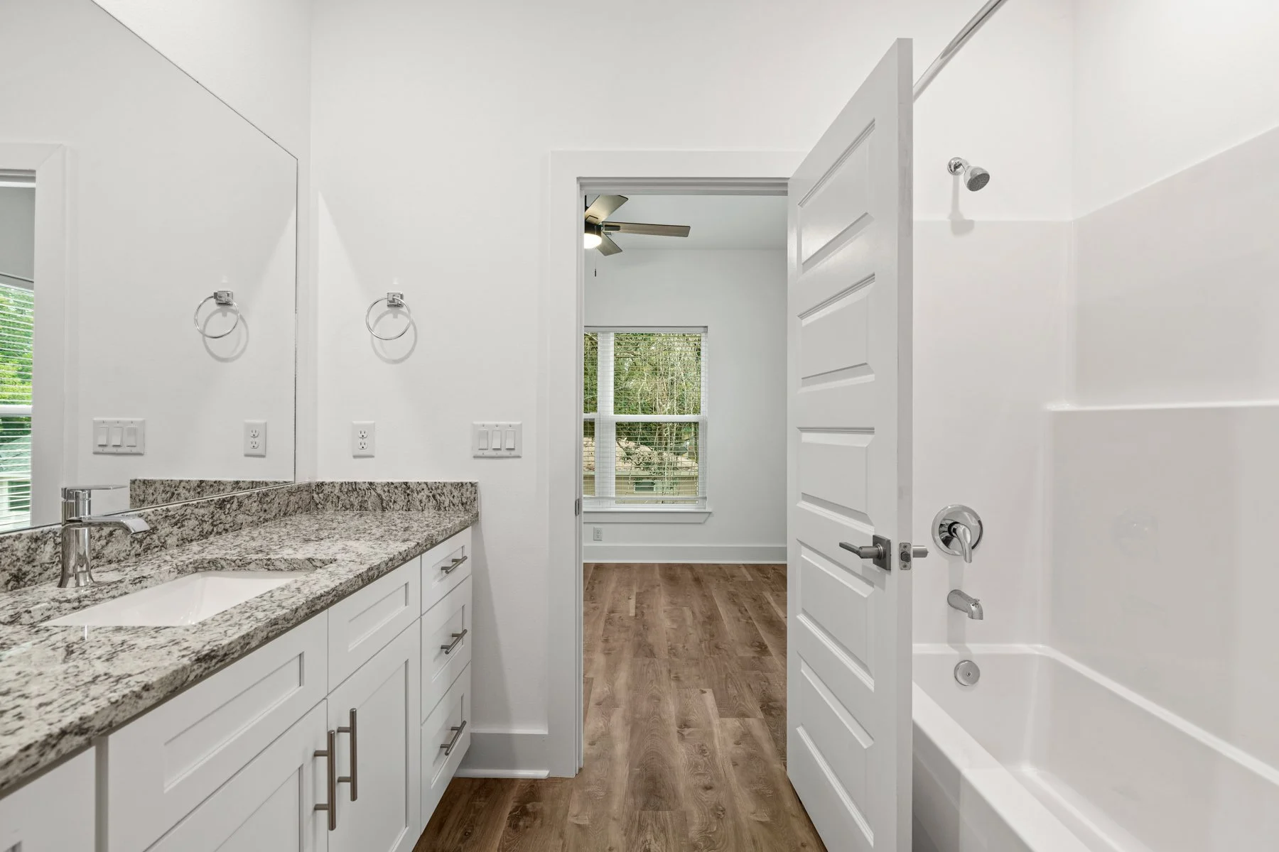 Bathroom with granite countertop vanity, white cabinets, wall mirror, towel rings, electrical outlets, a window with blinds, wooden flooring, and a walk-in shower with a door.