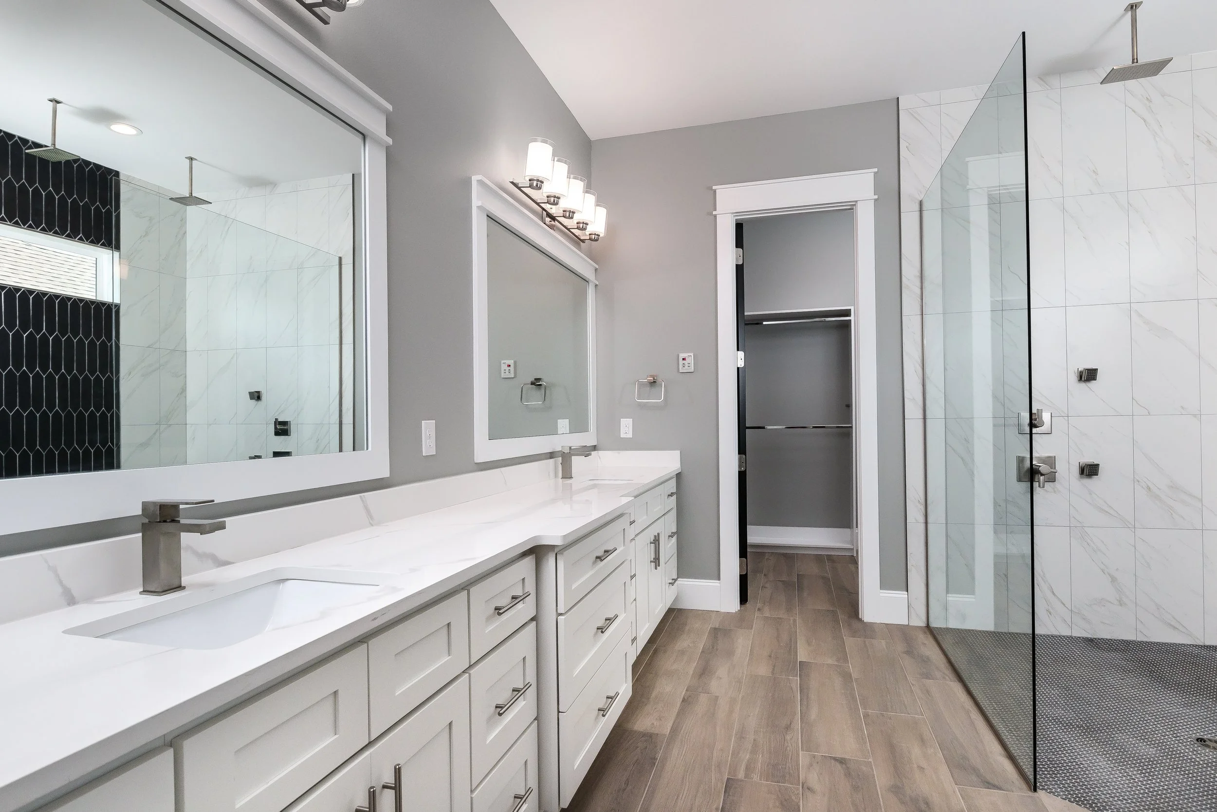 Modern bathroom featuring a double sink vanity with white cabinets, large mirrors, gray walls, a glass-enclosed walk-in shower with white marble tiles, and wood-look flooring.