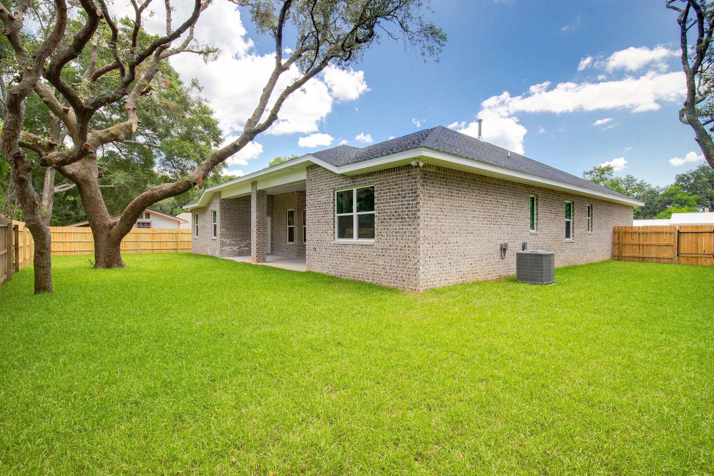 Backyard with a well-maintained grassy lawn, a large tree with sprawling branches, a brick house with multiple windows, a wooden fence, and a cloudy blue sky.