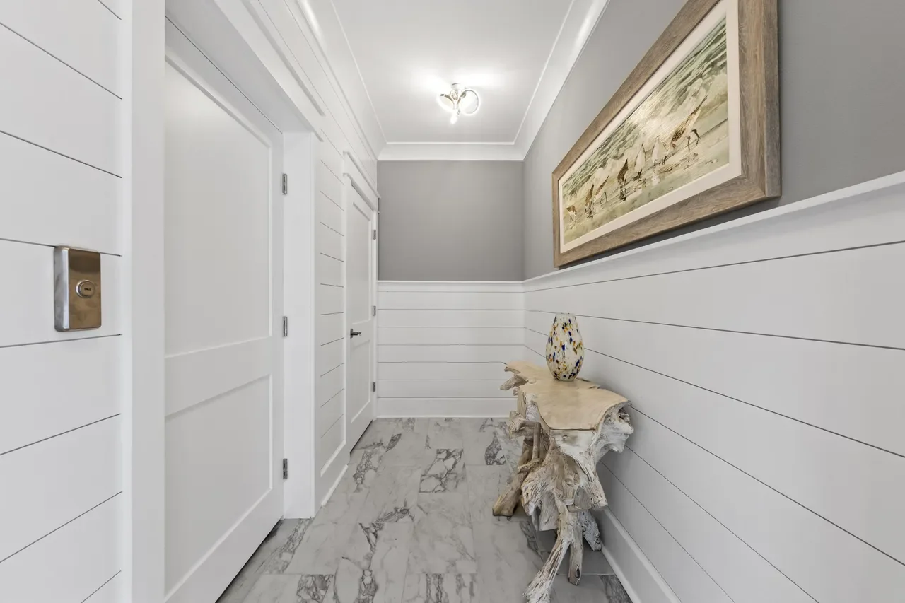 A hallway with gray walls, white shiplap paneling, and a marble tile floor. There are two white doors, a decorative table made from a piece of driftwood, a colorful ceramic vase, a large framed artwork of sailboats on water, and a ceiling light fixture.