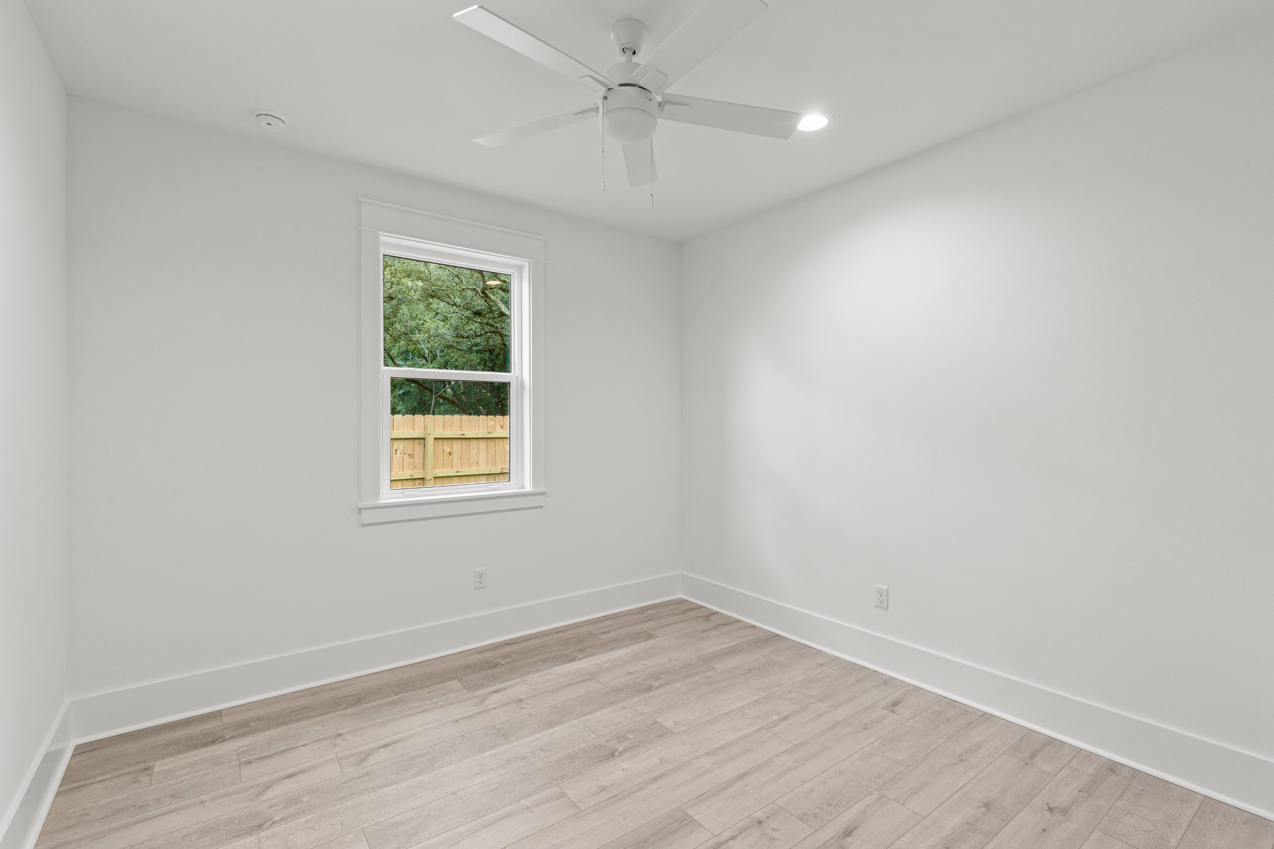 Empty white room with a window showing trees and a wooden fence outside, ceiling fan, and light wood flooring.