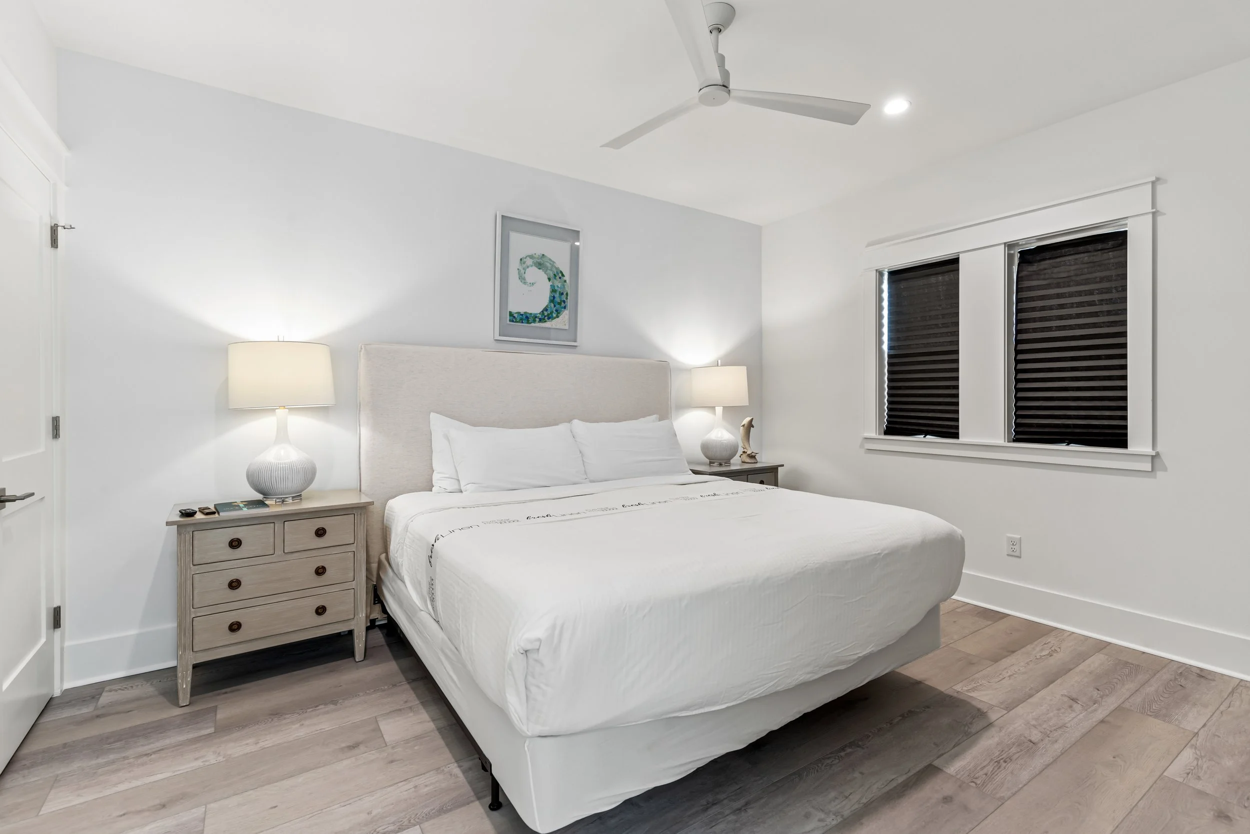 A bedroom with a white bed, bedside tables, lamps, dark window blinds, and a framed artwork of a seahorse on the wall.