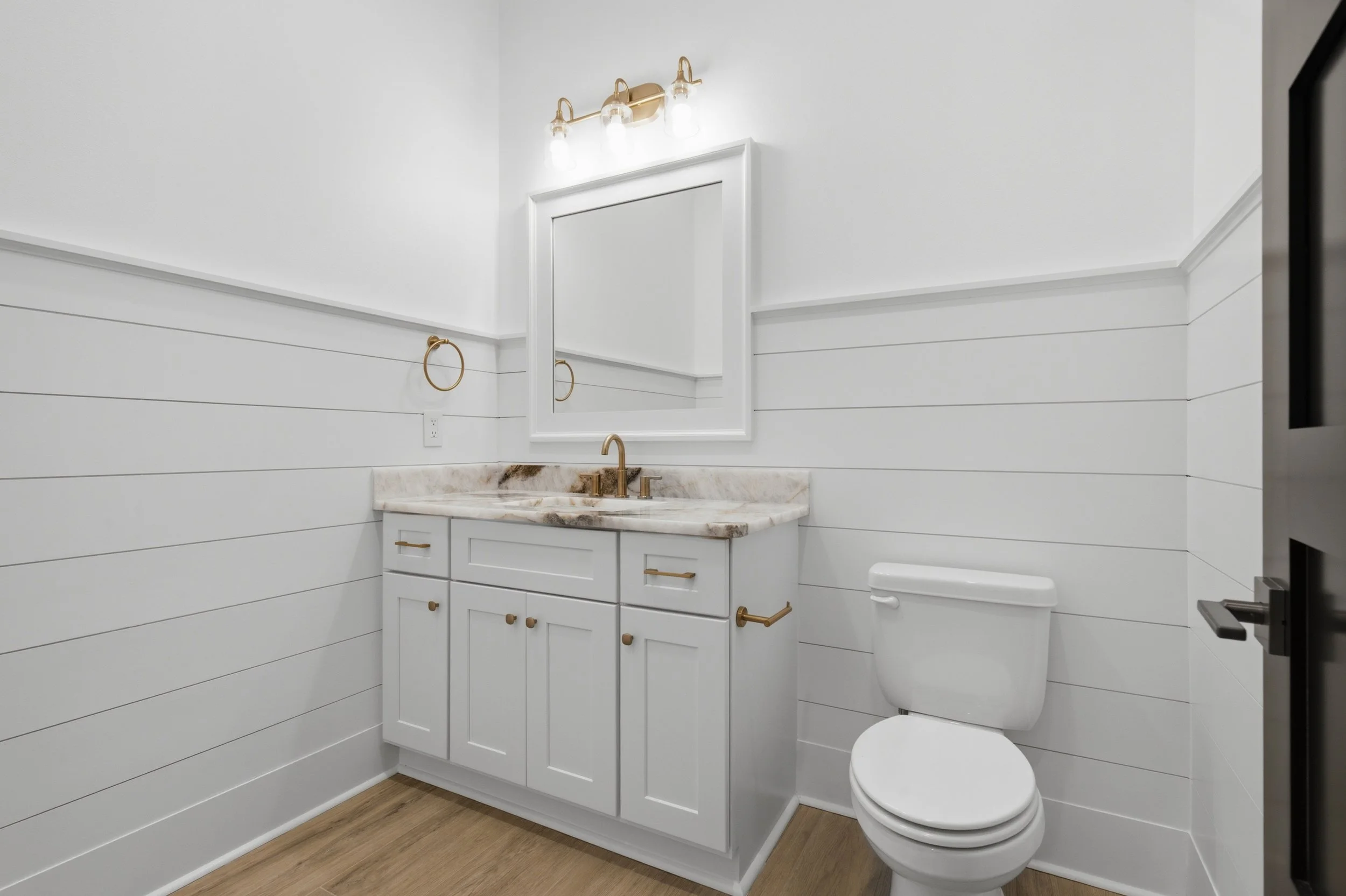A modern bathroom with white shiplap walls, a white vanity with a marble countertop, a gold faucet, a large mirror, a mounted light fixture with three lights, a towel ring, a toilet, and hardwood flooring.