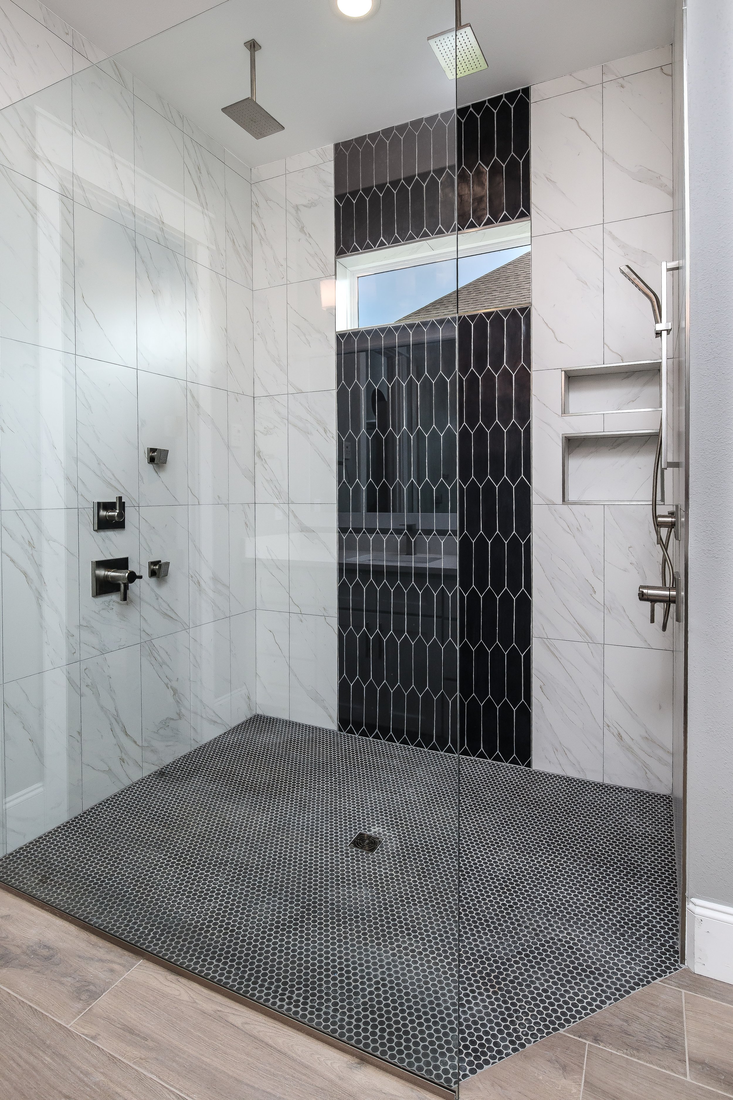 Modern walk-in shower with white marble walls, black patterned accent wall, black hexagonal floor tiles, and stainless steel fixtures including a rainfall showerhead and a handheld shower head. The shower has a glass enclosure and a window showing a nearby roof and sky.