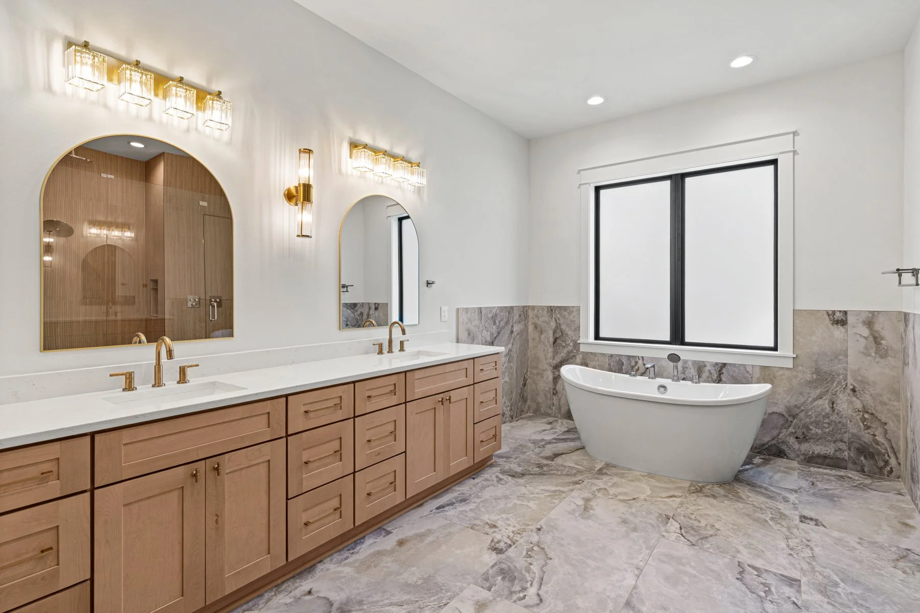 Modern bathroom with double vanity, wooden cabinets, white countertop, gold fixtures, large mirrors, wall-mounted lights, soaking tub near window, gray marble tile floor and walls, white walls.