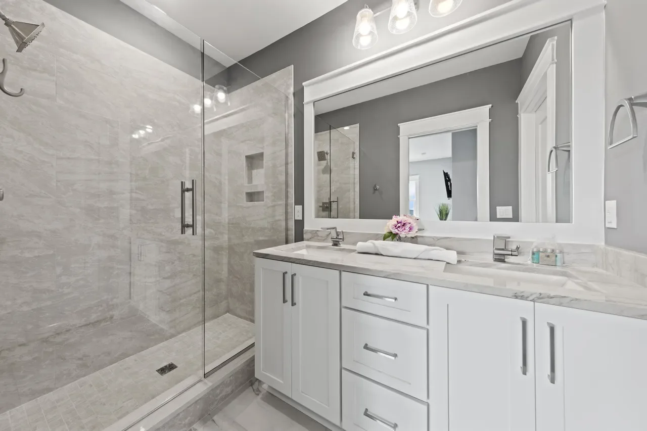 Modern bathroom with a glass-enclosed shower, white vanity with a marble countertop, large mirror, and gray walls. Decor includes a flower on the counter and small toiletries.