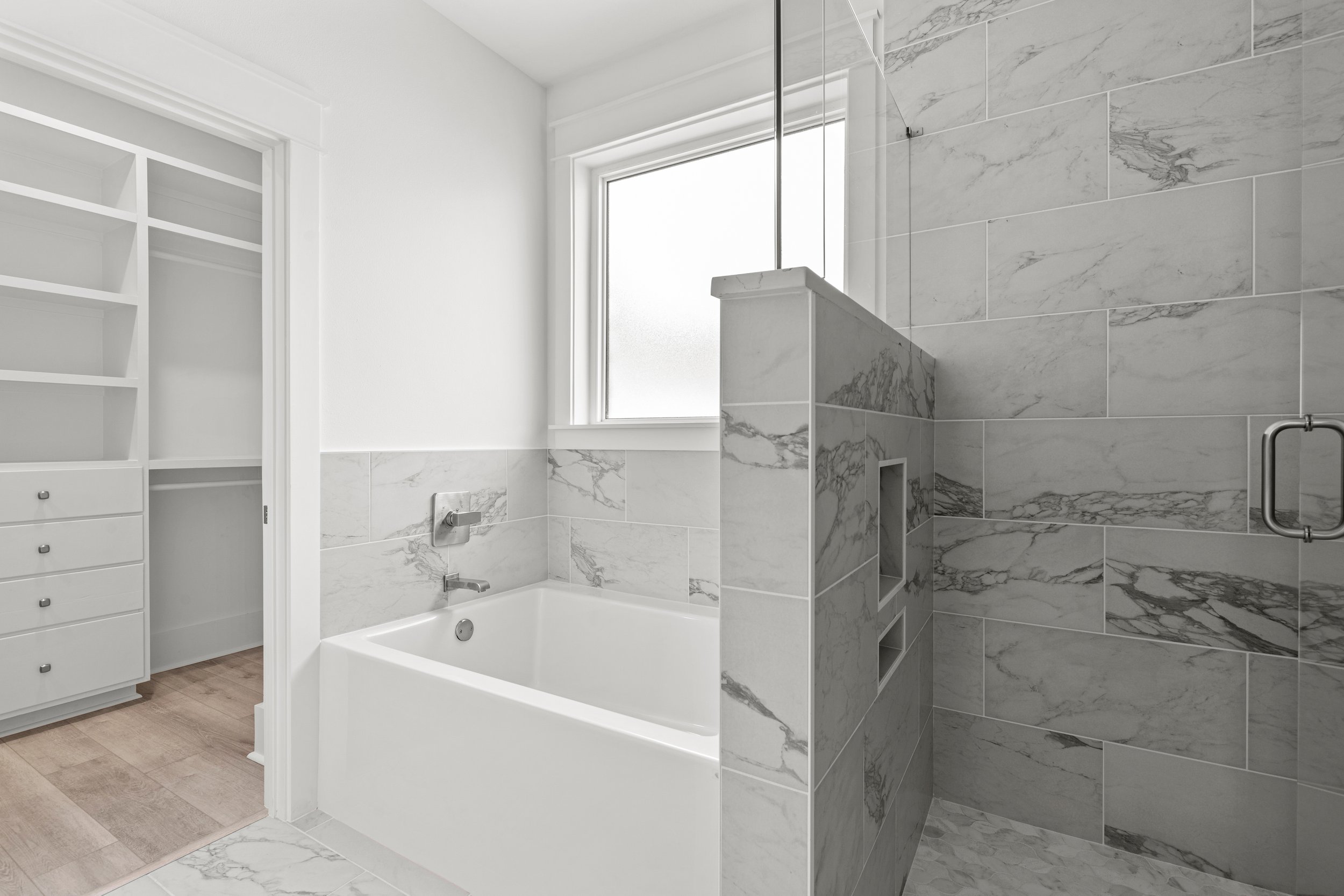 Modern bathroom with white walls, marble tiles, bathtub, window, and a built-in shower with glass door.