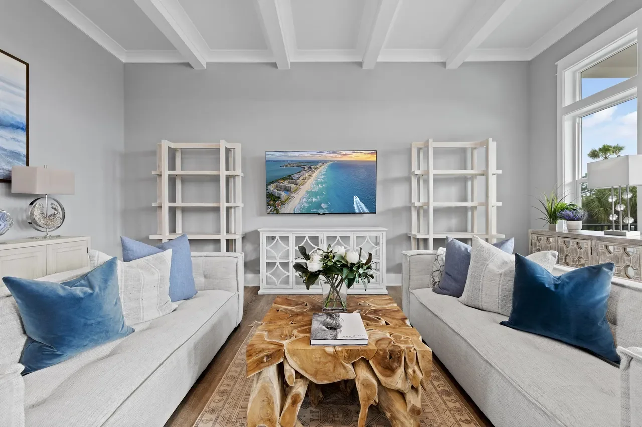 Living room with two white sofas with blue and white pillows, a wooden coffee table, a wall-mounted TV, white shelving units, a side board, and large windows showing a view of the sky and trees outside.