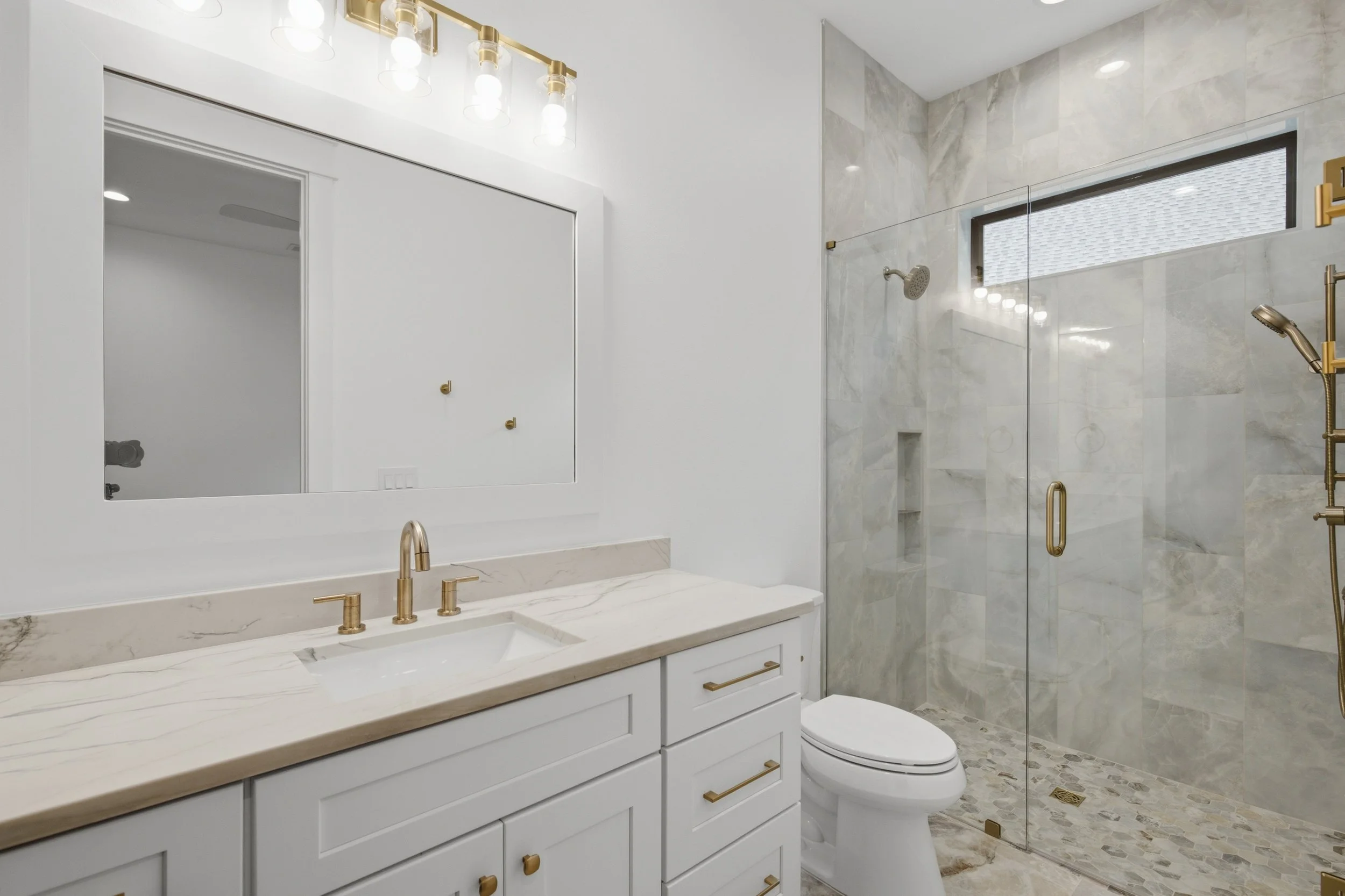 Modern bathroom with white vanity, marble countertop, gold fixtures, and a spacious glass-enclosed shower with marble walls and floor.