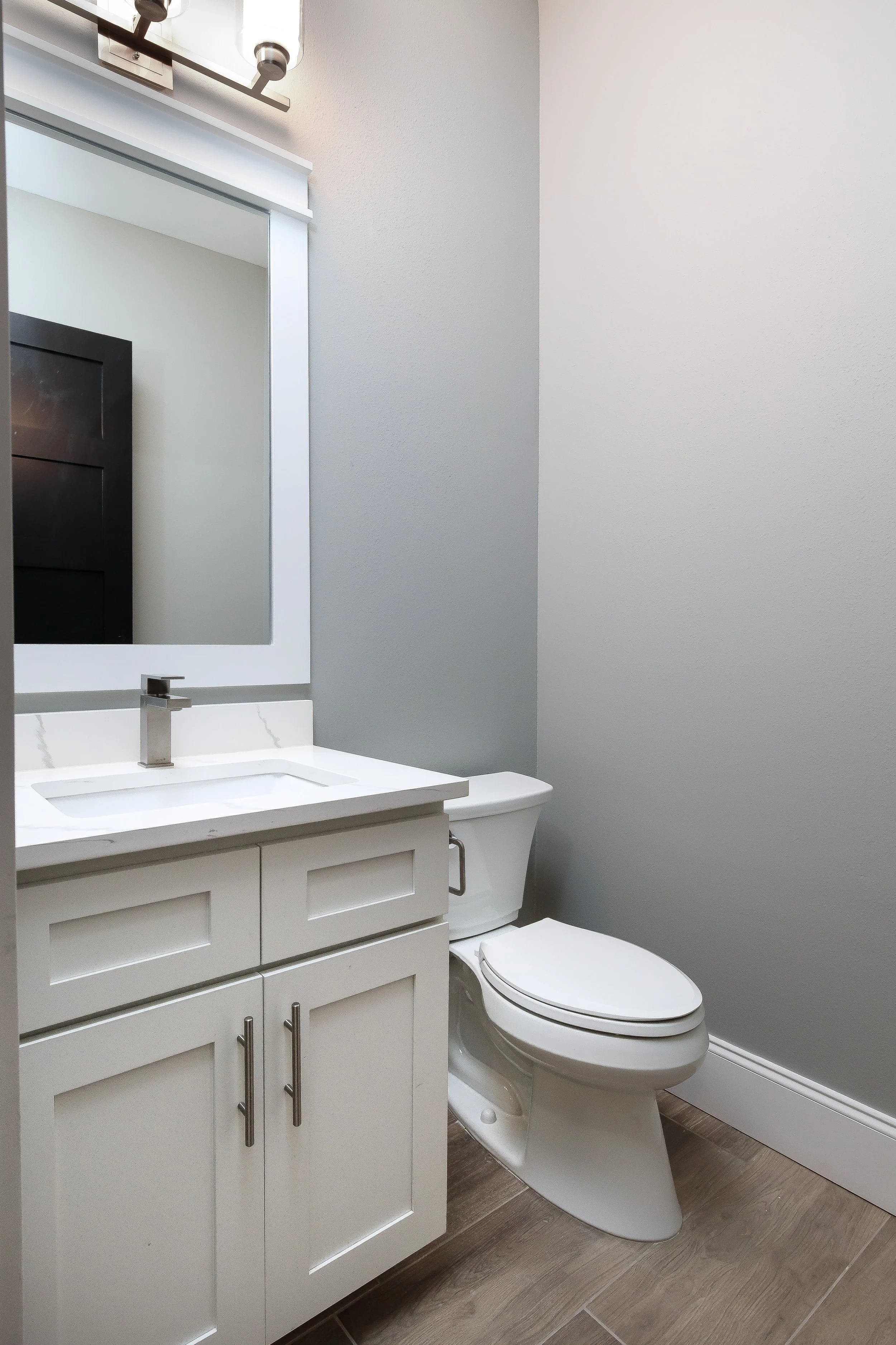 A modern bathroom featuring a white vanity with cabinetry, a sink with a chrome faucet, a large mirror with a light fixture above, a grey painted wall, and a white toilet on a wood-patterned tile floor.