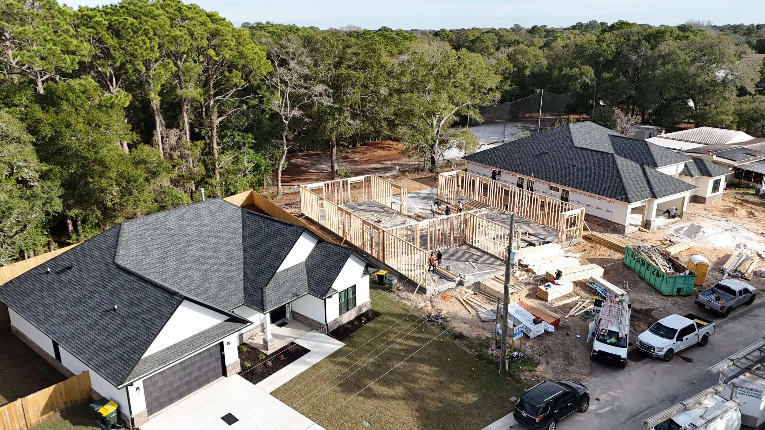 Aerial view of a construction site where a house is being built. The framing is in progress, surrounded by trees and neighboring houses with finished roofs. Construction equipment, workers, and materials are visible.