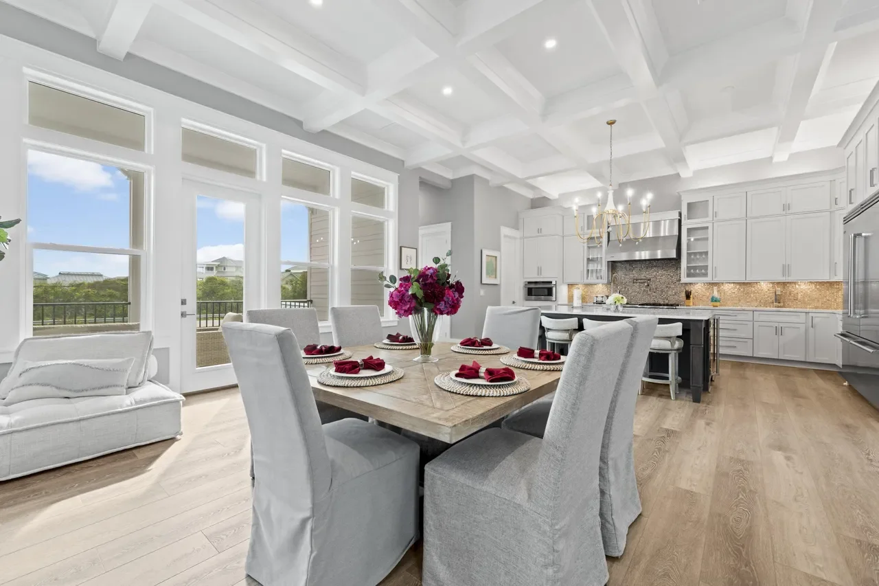 Bright, modern kitchen and dining area with large windows, white cabinetry, and a wooden dining table set with six plates and napkins, topped with a vase of pink flowers.