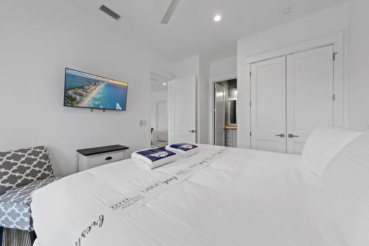 A bedroom with white walls, a large bed with white linens, a black nightstand, a patterned chair, and a mounted flat-screen TV showing an aerial view of a beach. There are white closet doors, a bathroom door, and a view into a bathroom with a sink. The room has ceiling lights and a vent.
