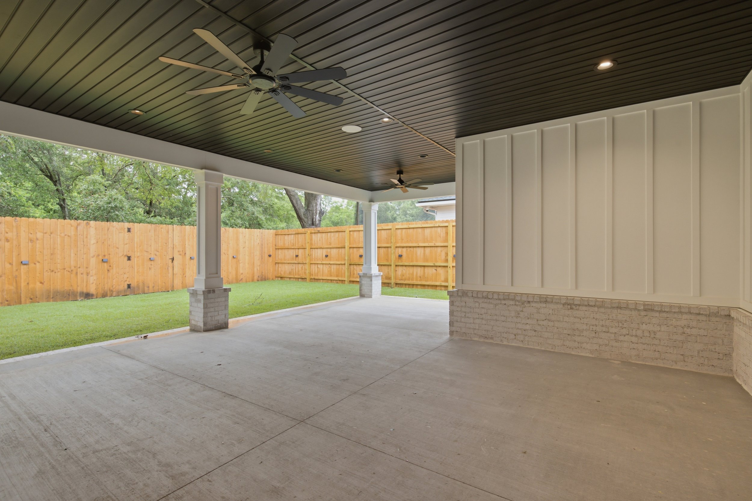 Covered patio area with concrete flooring, ceiling fans, and recessed lighting, overlooking a backyard with a grass yard and wooden fence, and trees in the background.