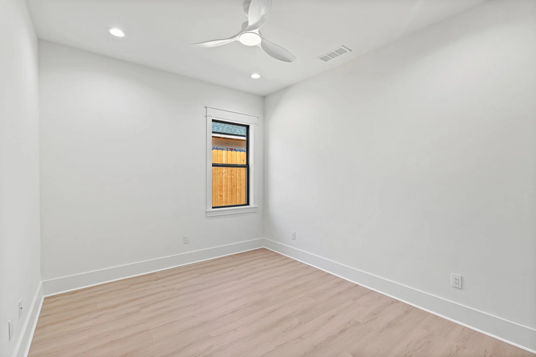 Empty room with white walls, a window showing a wooden privacy fence outside, a ceiling fan, and light hardwood floors.