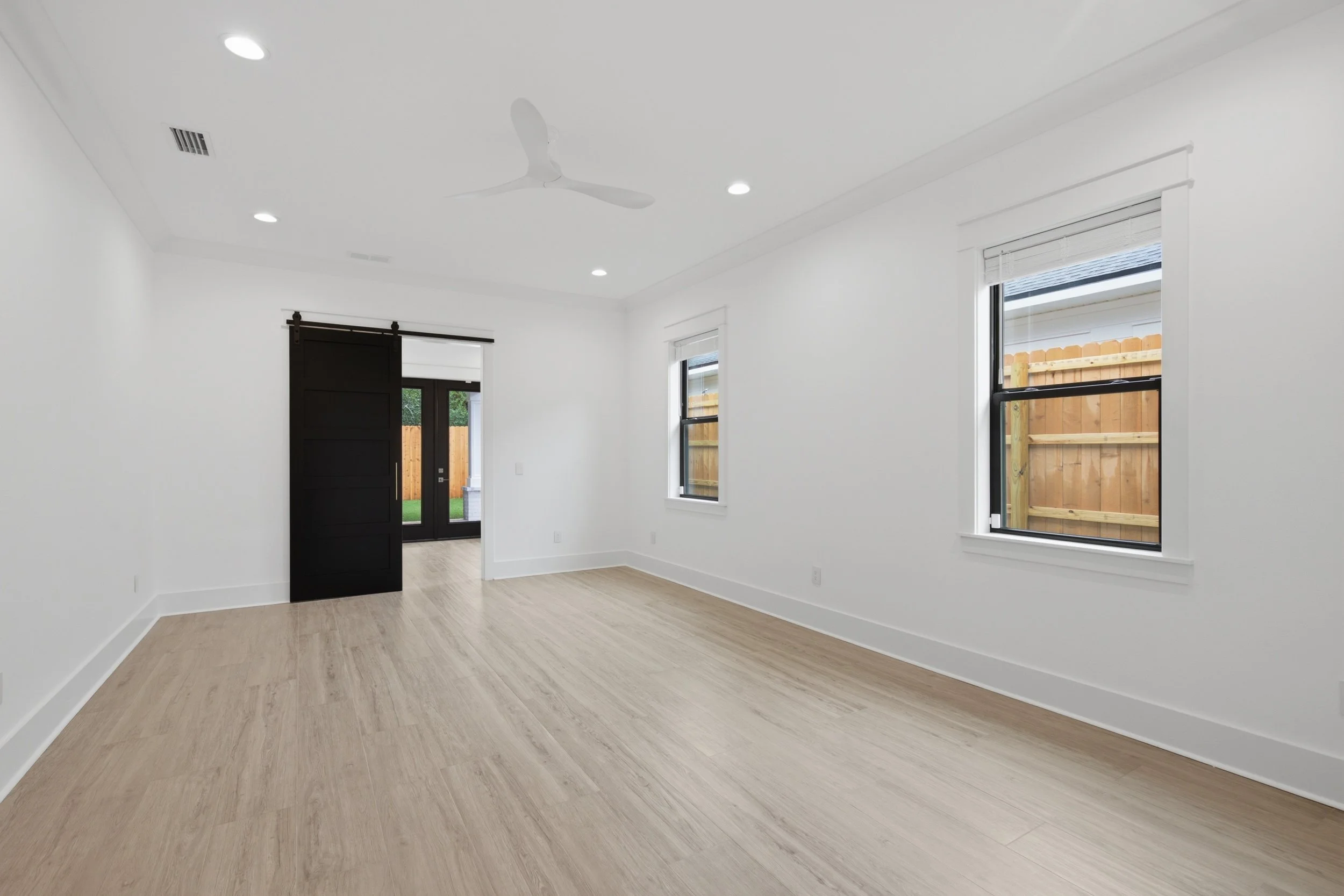 Interior view of a bright, empty room with white walls, light wood flooring, windows with white blinds, a black door, and a ceiling fan.