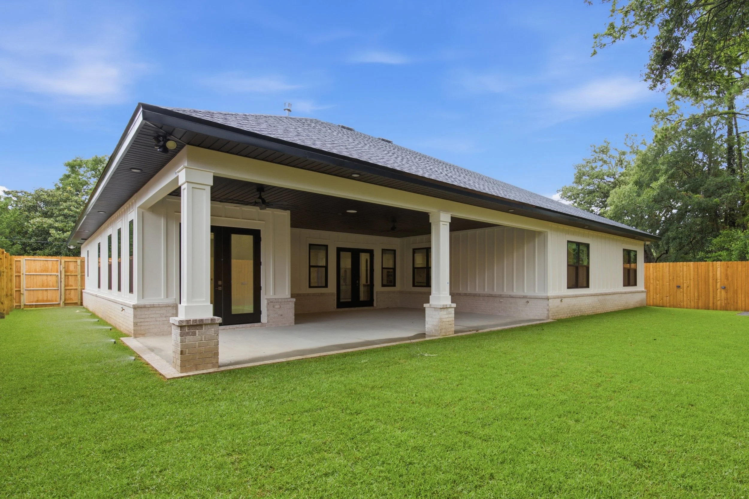 Backyard with a covered patio, white exterior walls, black-framed glass doors, brick columns, and a green lawn under a blue sky with trees.