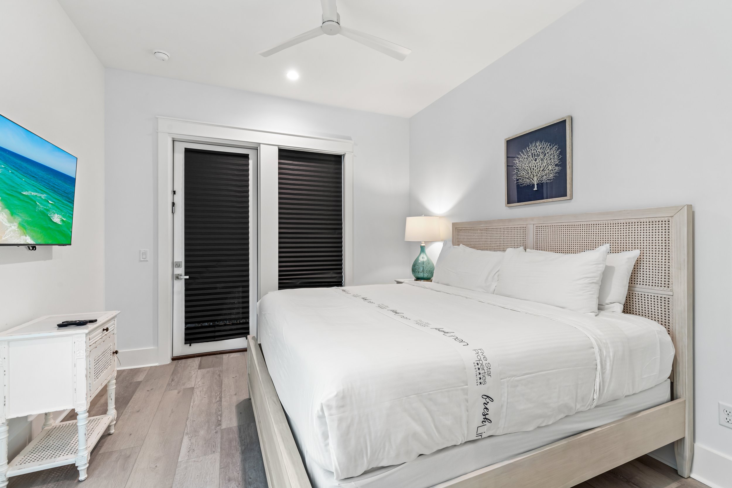 A bedroom with a large bed, white linens, and a woven headboard. There is a nightstand with a decorative glass lamp, a flat-screen TV on the wall, and a closet with black blinds. The room has white walls, a ceiling fan, and a framed art piece of a white tree on a dark background.