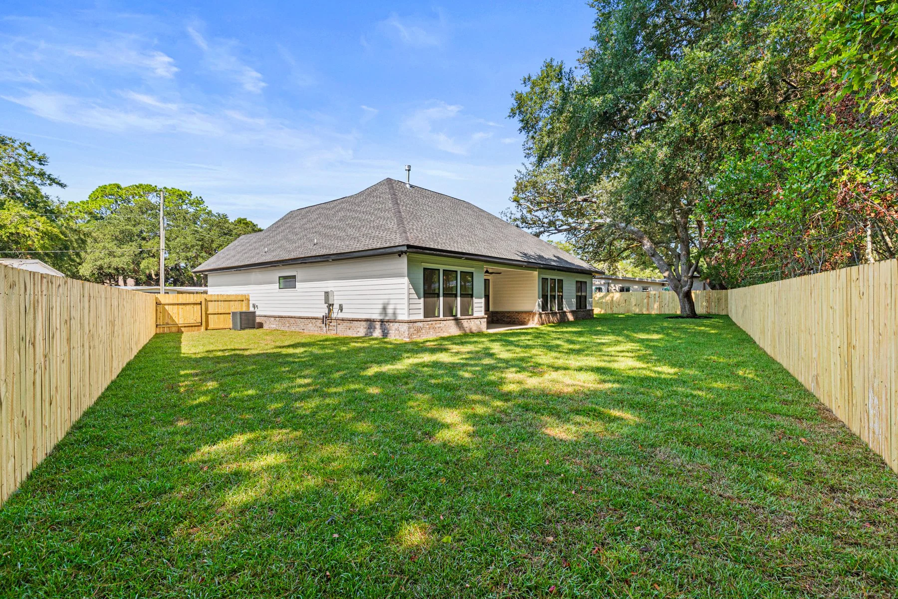 Backyard with green grass, wooden fence, large trees, and a white house with a dark roof under a clear blue sky.
