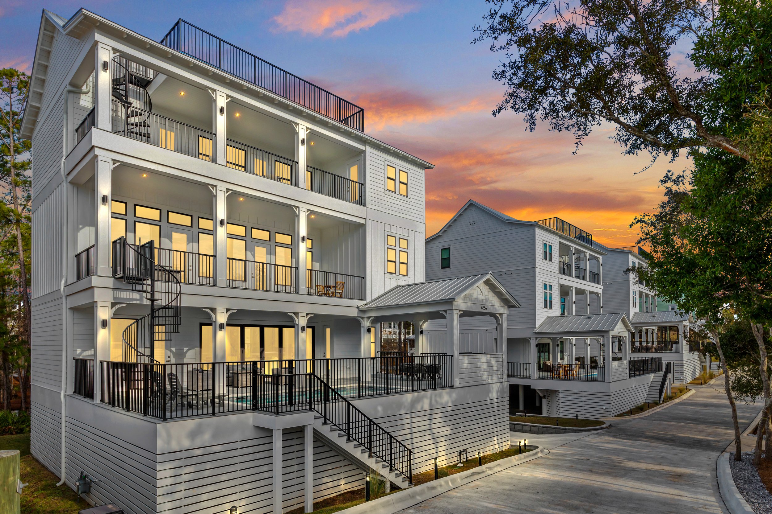 A modern white multi-story residential building with black railings and spiral staircase on the balconies, large windows glowing with interior light, surrounded by trees and a paved walkway, under a colorful sunset sky.