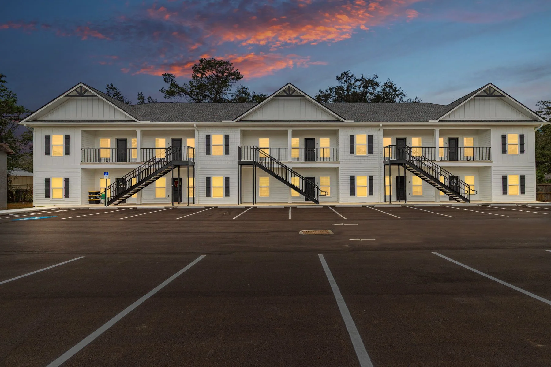 A two-story white apartment building with black shutters and exterior staircases, illuminated from within, against a sunset sky with a mostly empty parking lot in front.