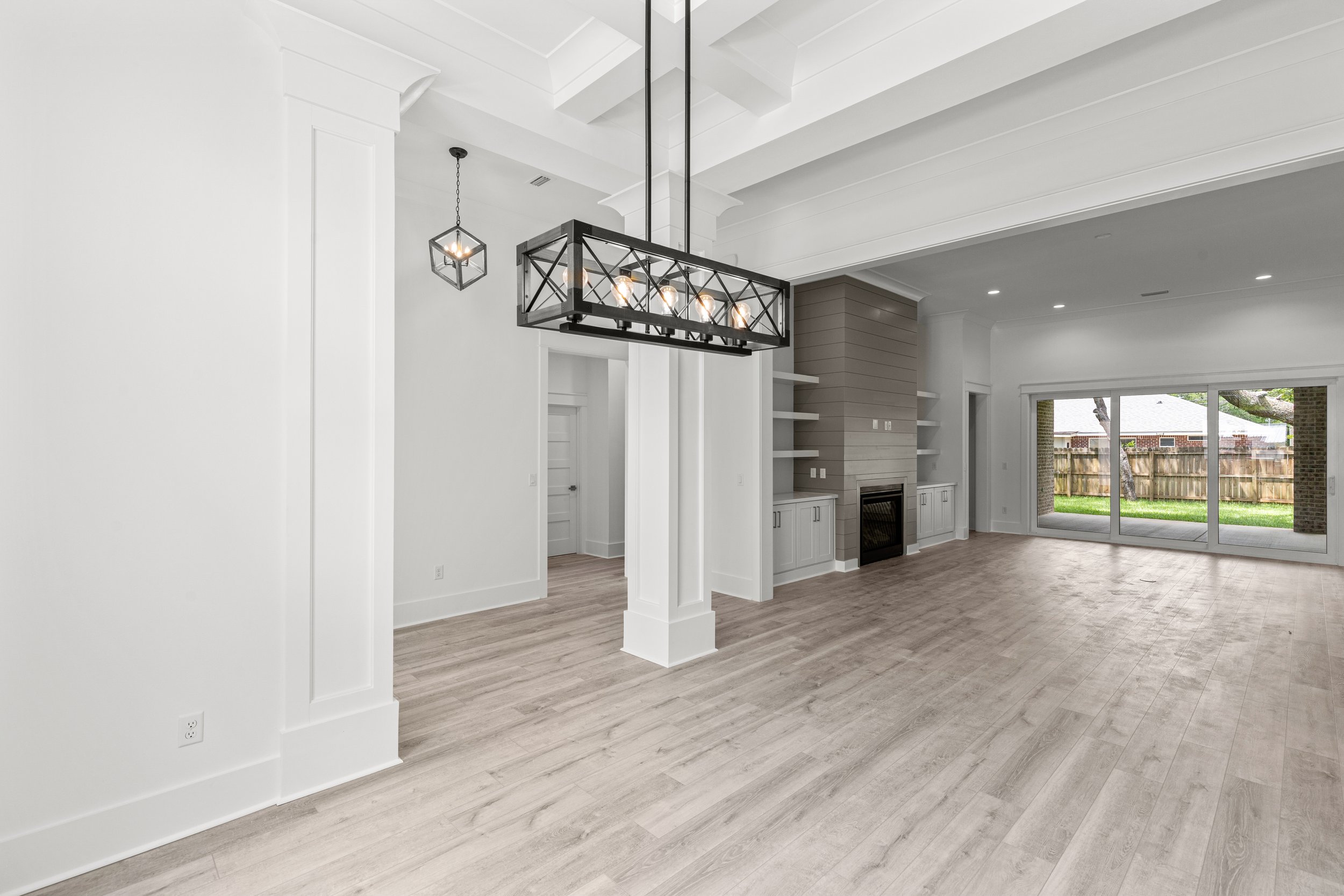 Open living room with white walls, vaulted ceiling, modern chandelier, built-in shelving, fireplace, large sliding glass door leading to backyard, light wood flooring.