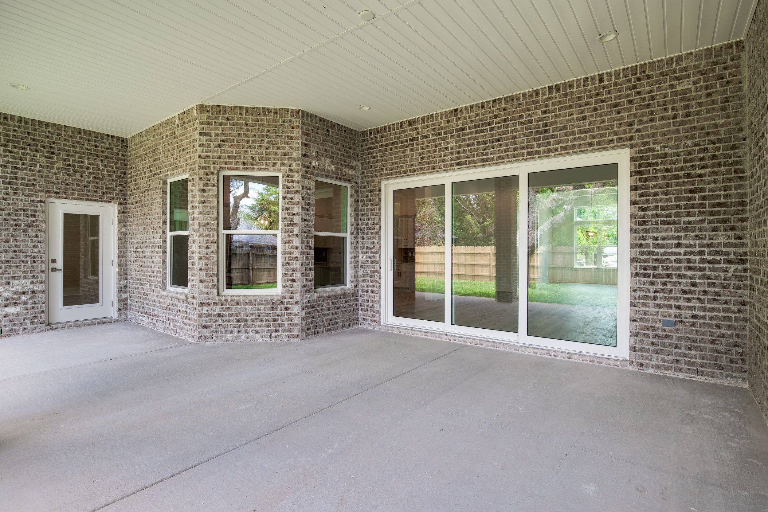 Covered outdoor patio with brick walls, large sliding glass door, and windows, with a concrete floor and a white ceiling.