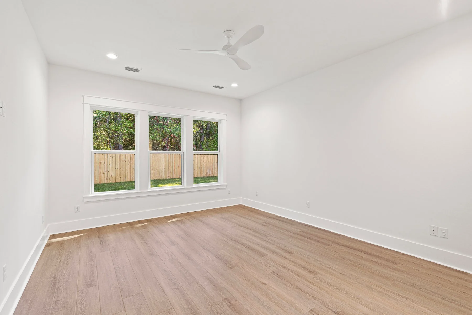 Empty room with white walls, wooden flooring, large window showing a backyard with a wooden fence and green trees, ceiling fan, and recessed lighting.