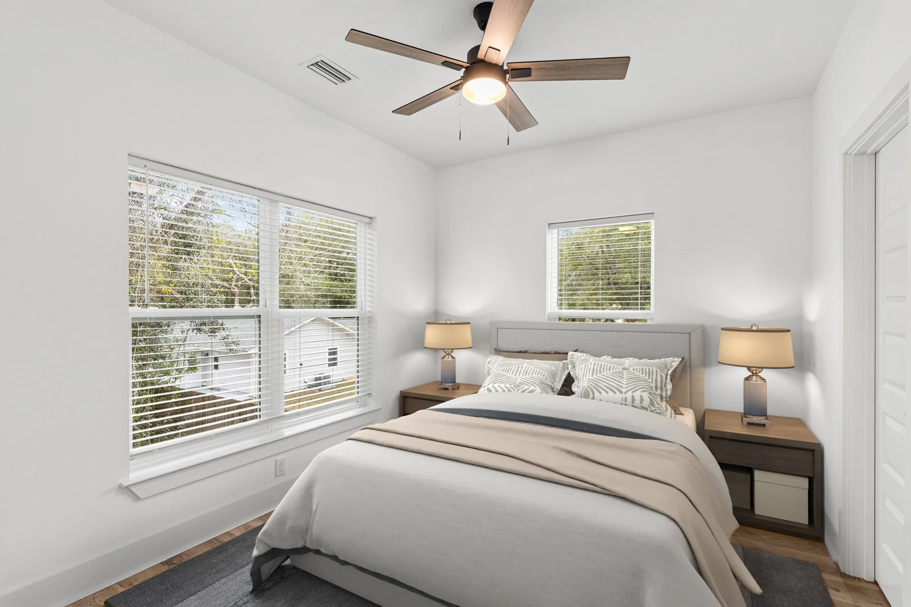 A cozy bedroom with a bed, two bedside tables with lamps, two large windows with blinds showing trees outside, and a ceiling fan with lights.