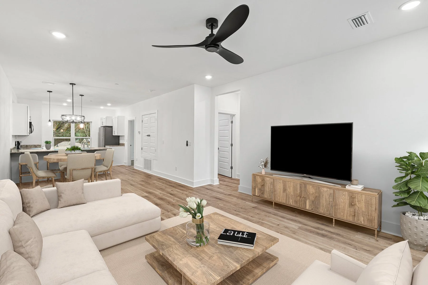 Modern living room with white walls, light beige couches, a wooden coffee table with a vase of white tulips and books, a TV on a wooden TV stand, a large green plant, and a dining area with a round table and eight chairs in the background. The space has wood flooring and is brightly lit with ceiling lights and pendant lights.