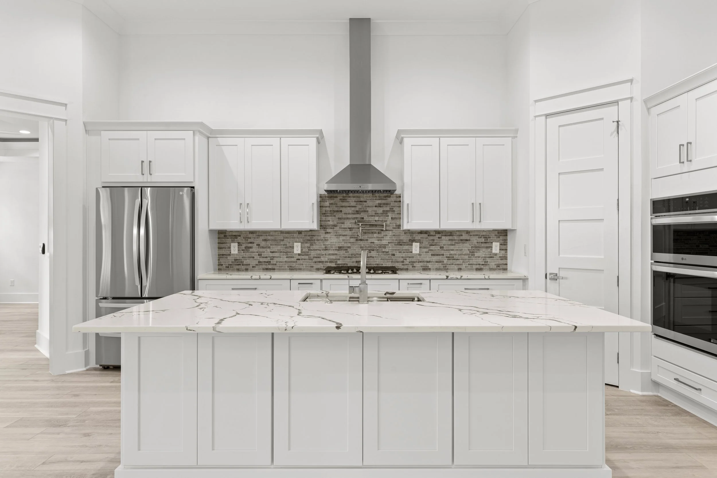 Modern kitchen with white cabinets, marble island, stainless steel refrigerator, and a brick backsplash behind the stove.