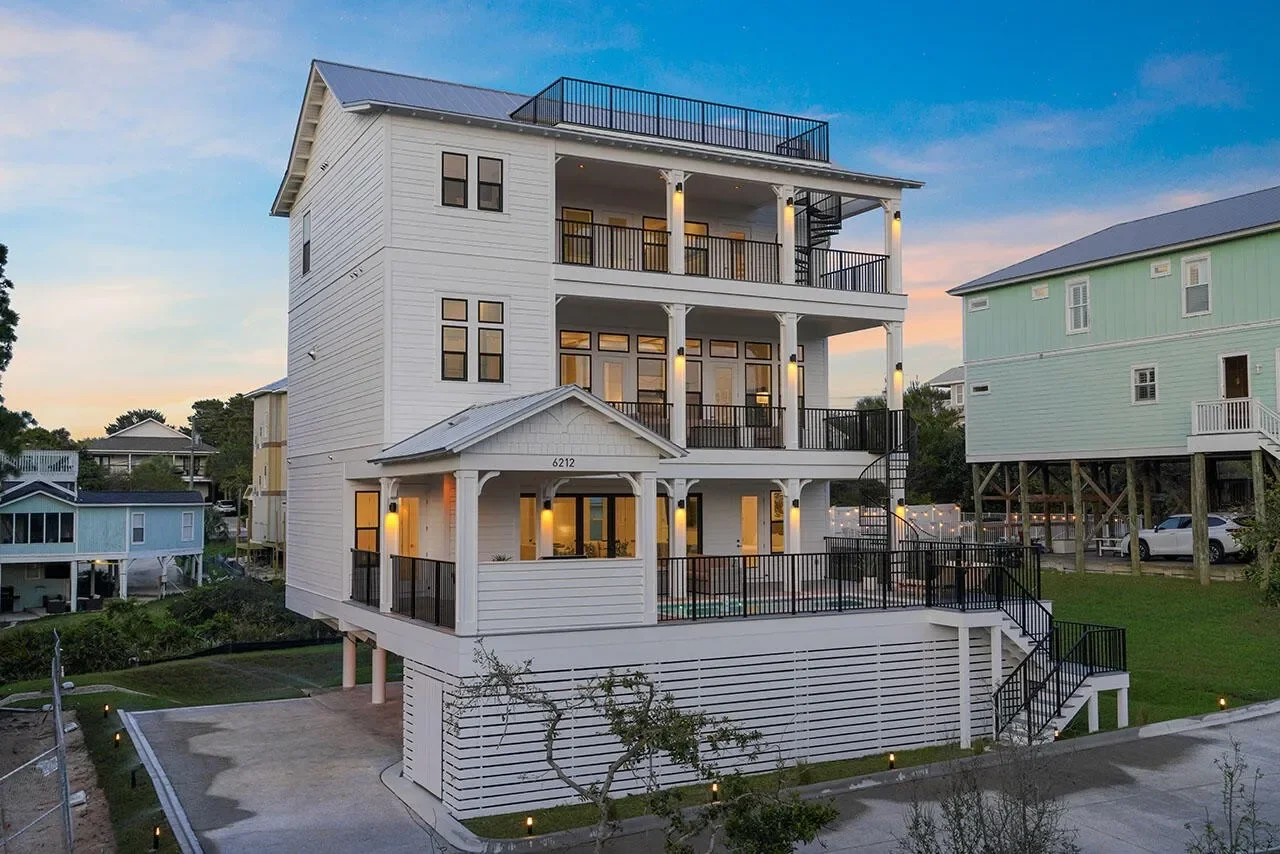 Multi-story white house with balconies and black railings, situated in a neighborhood at sunset.