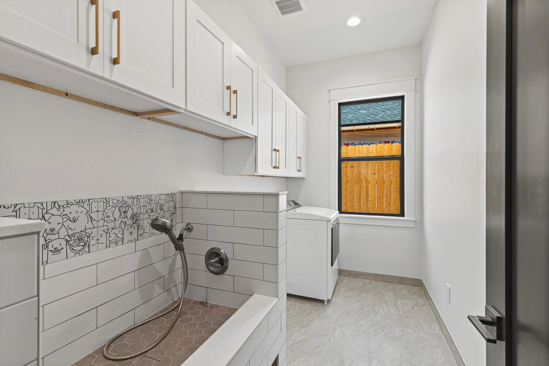 Laundry room with white cabinets, laundry washer, decorative animal face tiles, and a window overlooking a wooden fence.