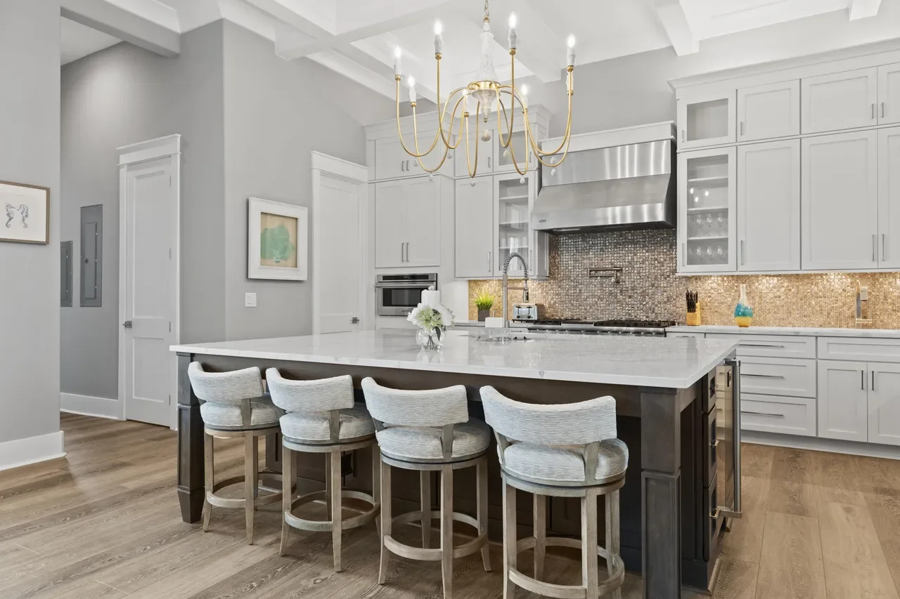 Modern kitchen with white cabinetry, a large kitchen island with a white marble countertop, four fabric-upholstered bar stools, a stainless steel range hood, and a gold chandelier.