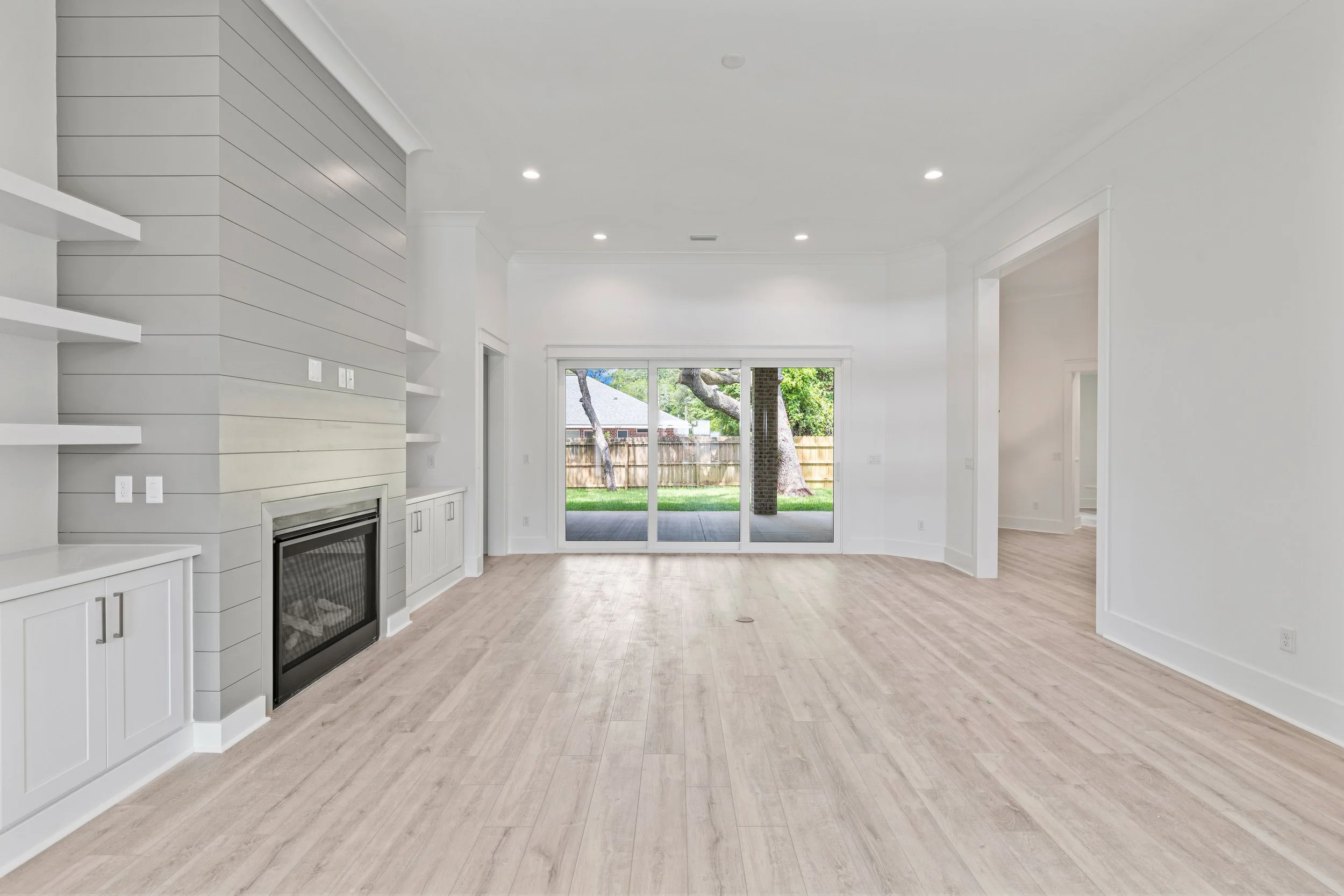 Empty living room with white walls and ceiling, light wood flooring, built-in white cabinets, a fireplace, and large sliding glass doors leading to a backyard with a tree and wooden fence.