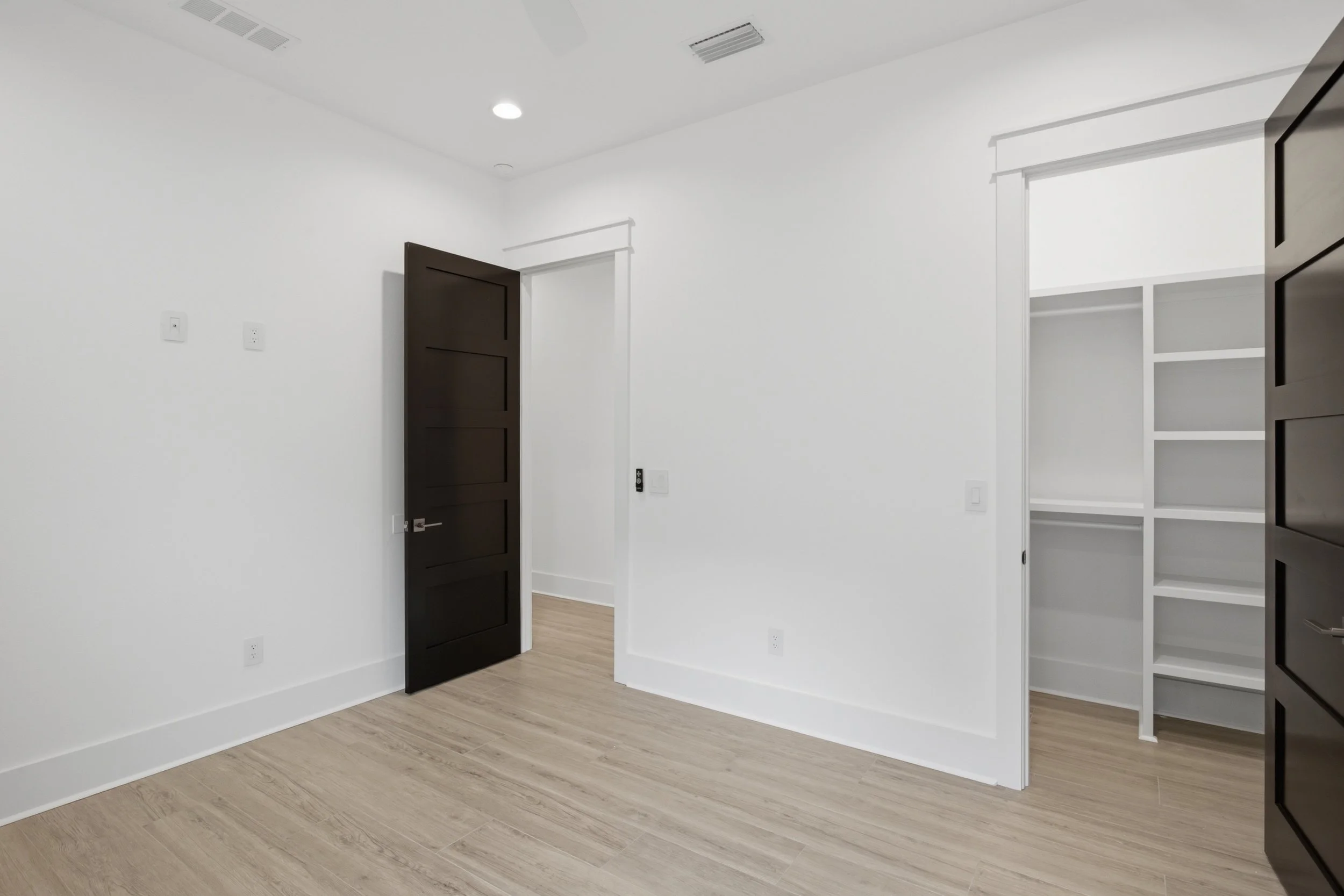 Empty room with white walls, a black door, built-in shelves, wooden flooring, and ceiling vents.