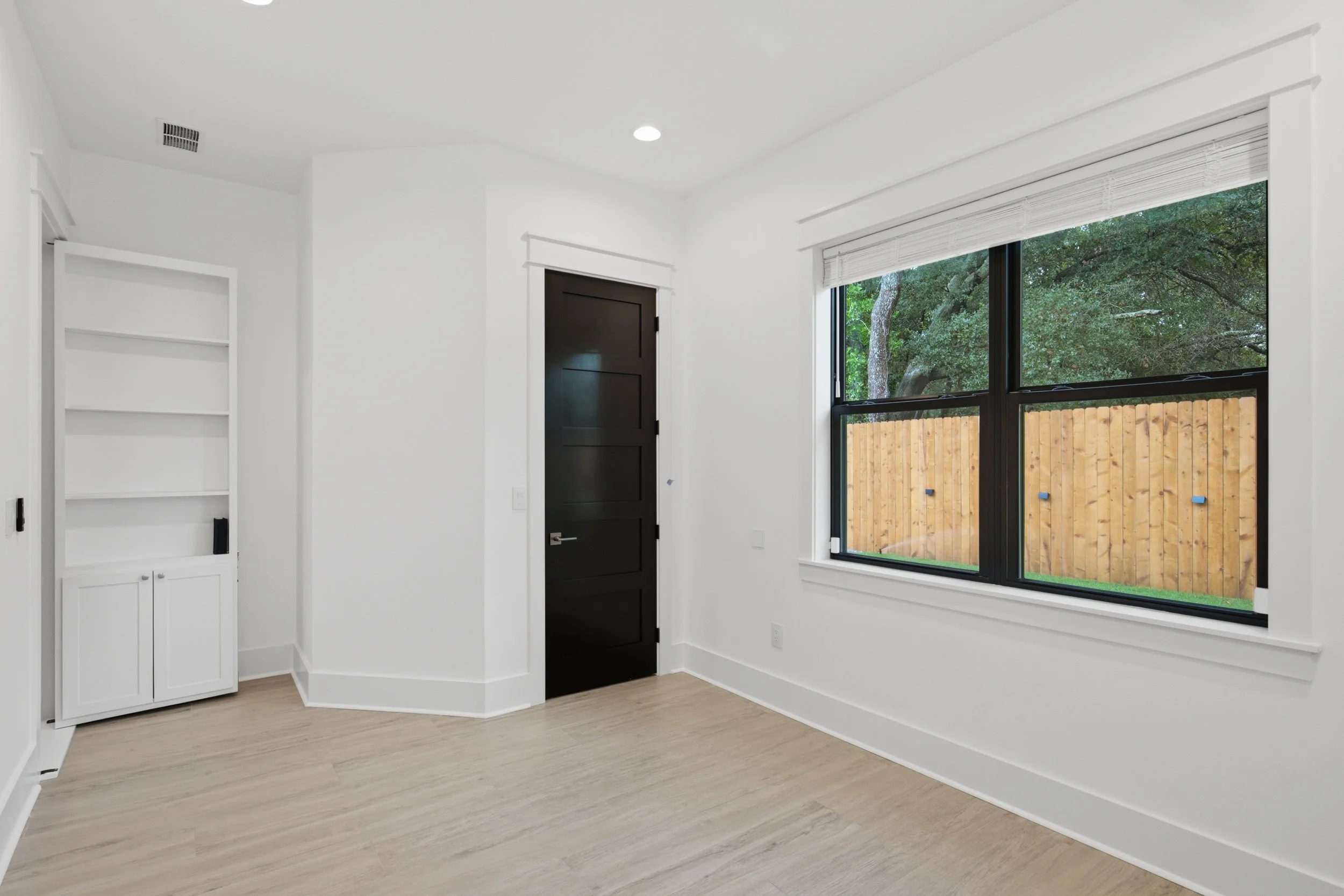 Empty room with white walls, a large window with blinds showing green trees outside, a black door, built-in white shelves, and light wooden floors.