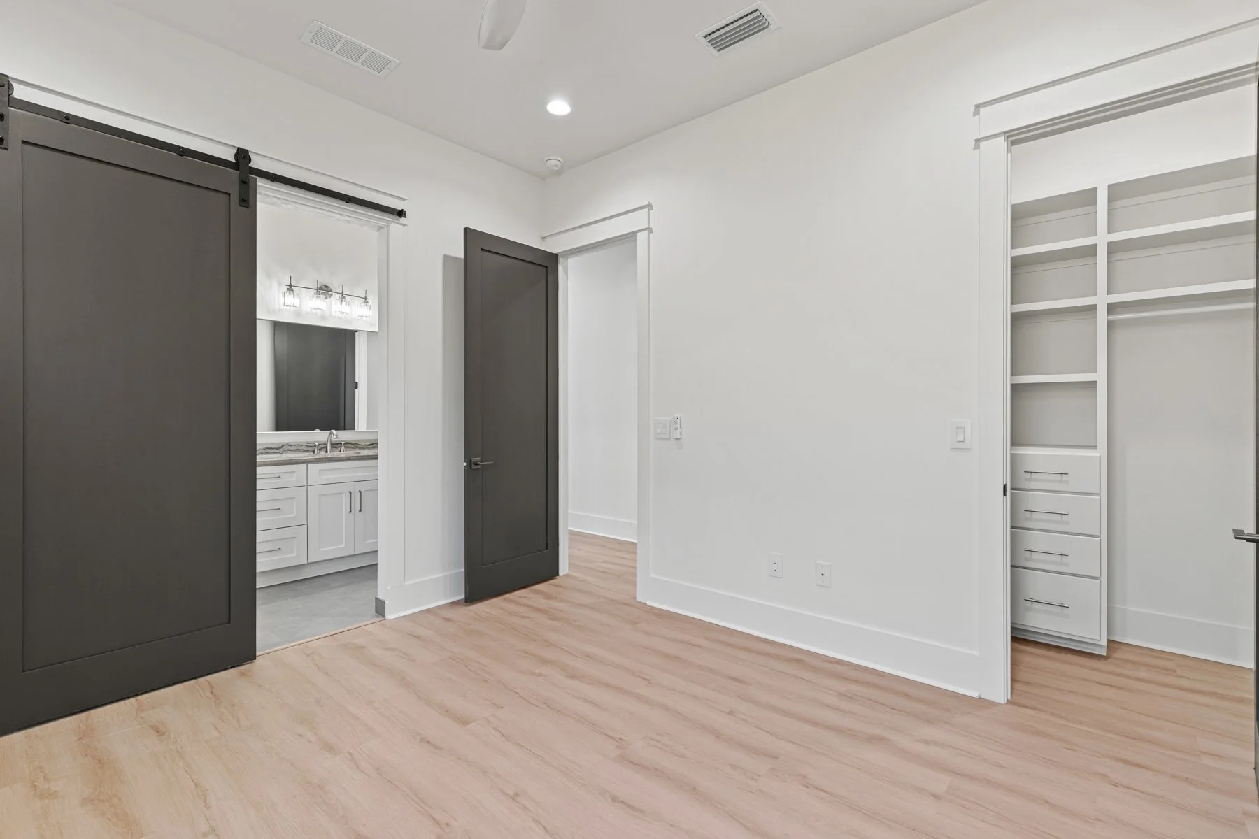 An empty room with white walls, light wood flooring, a sliding barn door, a closet with shelves, and a doorway leading to a bathroom with a vanity mirror and sink.