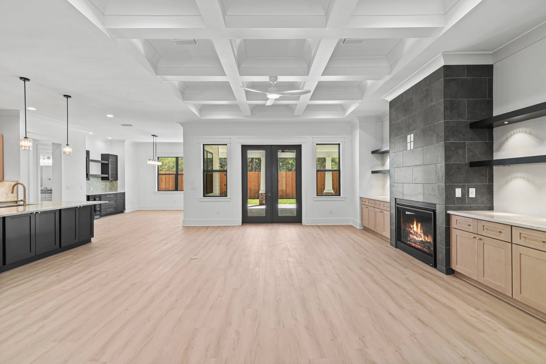 Open-concept living room and kitchen with modern design, light wood flooring, white walls, a gray stone fireplace, black and light wood cabinets, black double doors, large windows, and a ceiling fan.