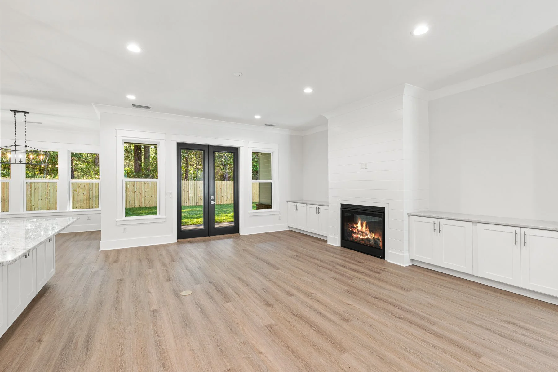 Bright, modern living room with white walls, wood flooring, a fireplace with a fire, white cabinetry, large windows, and black double doors leading outside to a grassy backyard.