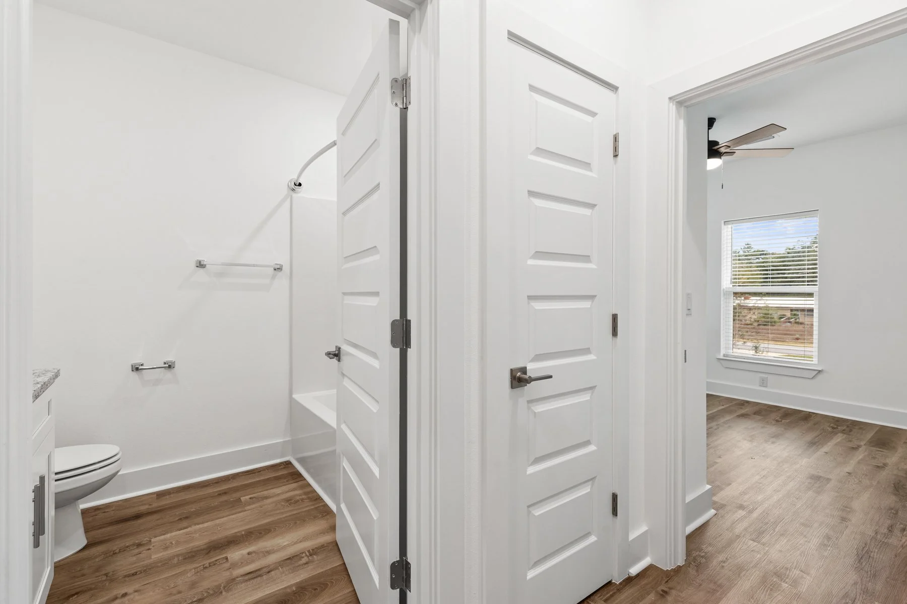 A bathroom with two white doors separating the toilet area from the bedroom. The toilet is visible on the left. The bedroom has a window with blinds, a ceiling fan, and wood flooring.