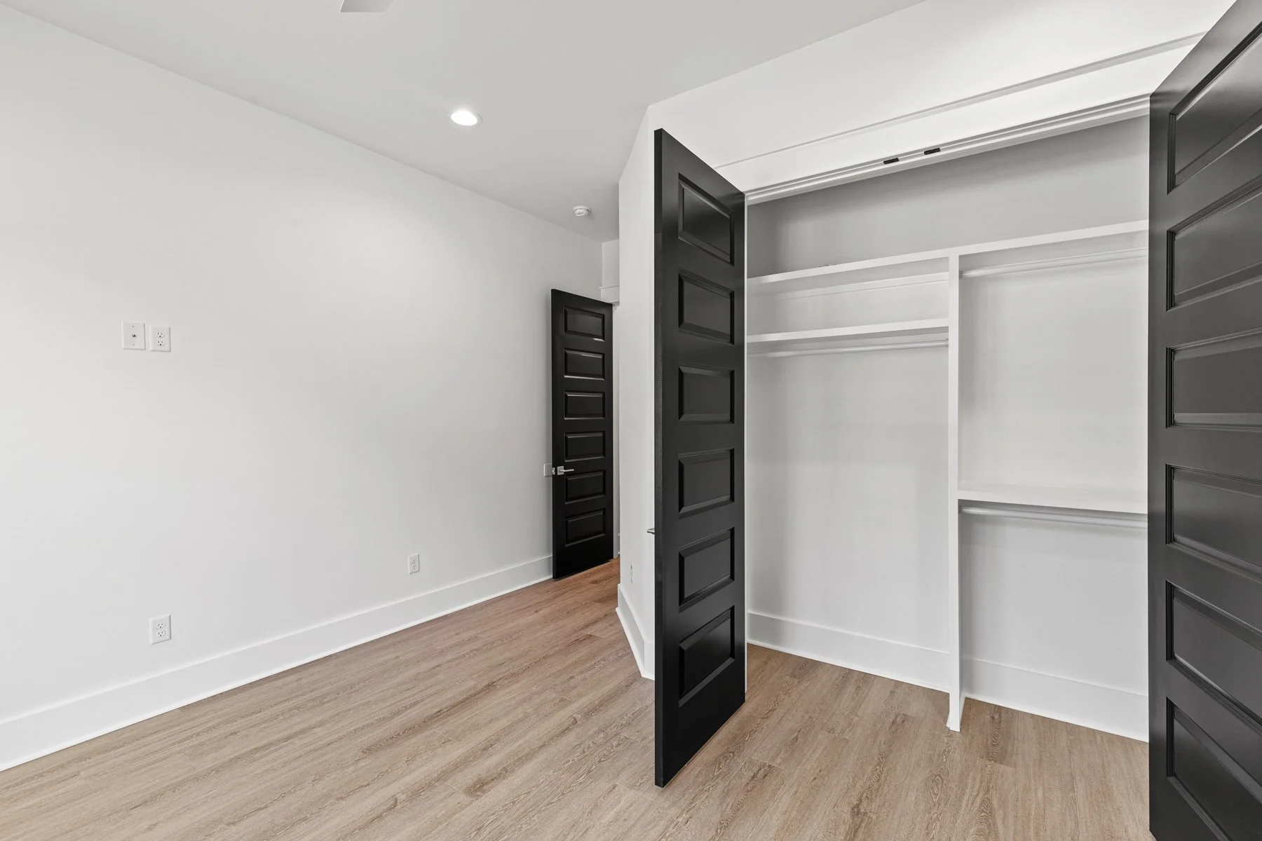 Empty room with black door, white walls, and open closet with white shelves and black doors, wood flooring, and ceiling light.