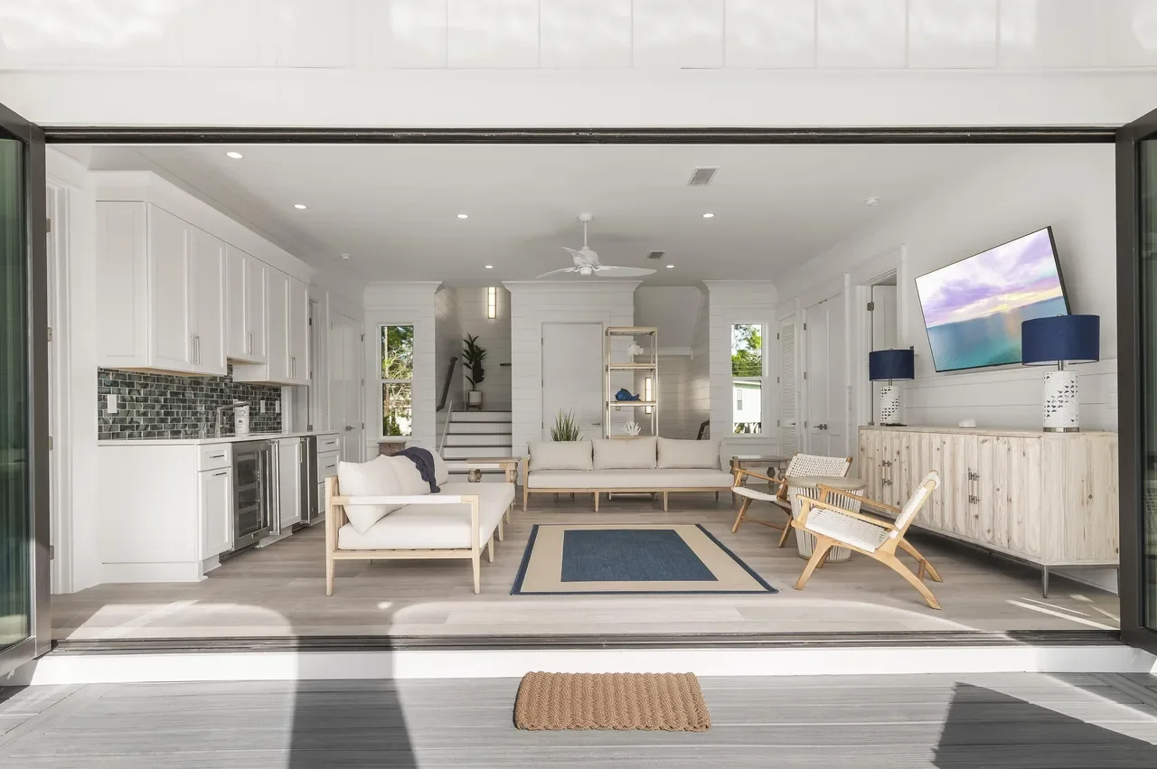 Bright, minimalistic living room with white walls, ceiling, and furniture, featuring a blue rug, wooden chairs, a sofa, and a mounted television with two blue lamps.