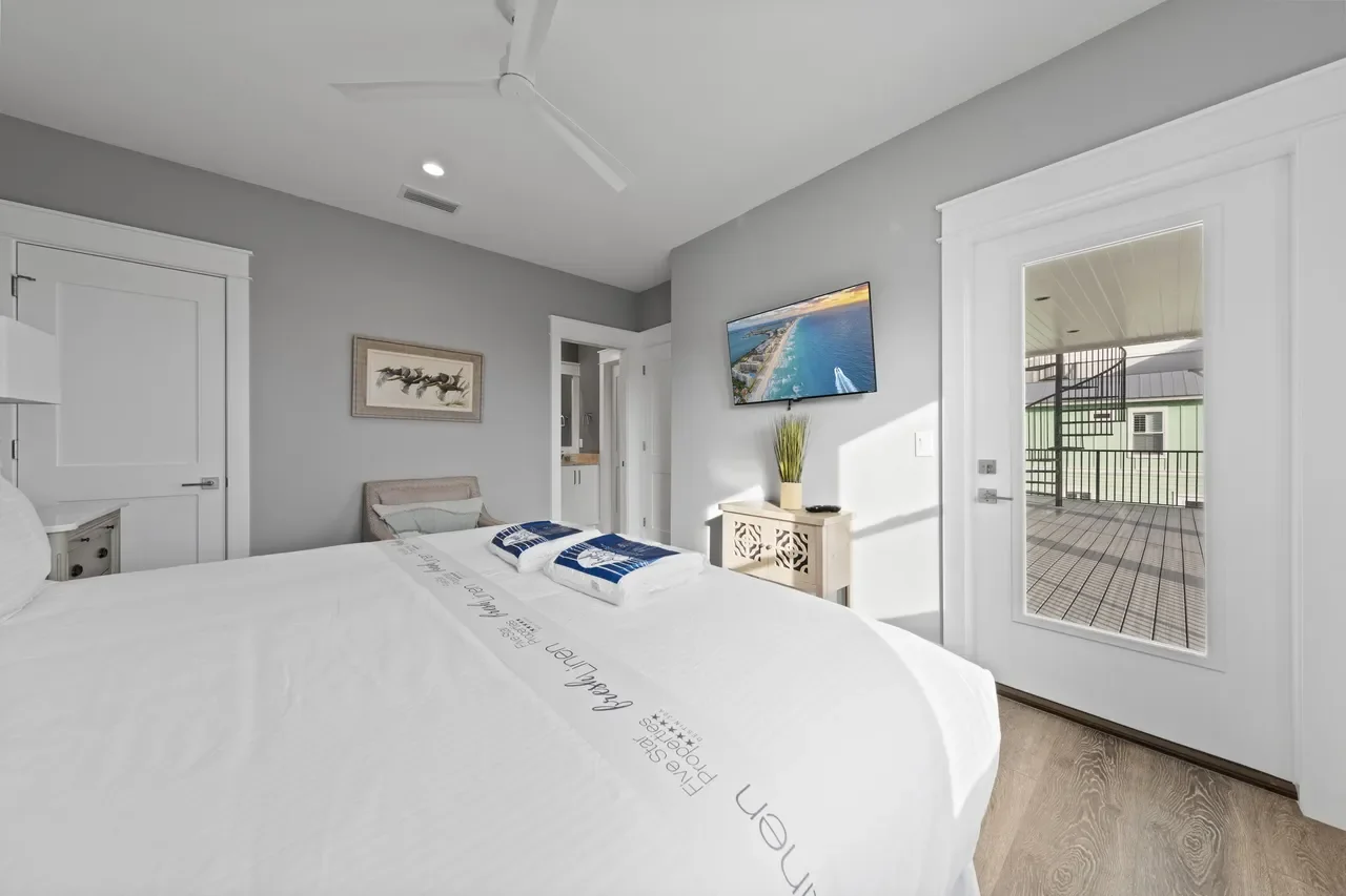 Bright hotel bedroom with a king-sized bed, white bedding, a wall-mounted TV, a small decorative table, and a sliding glass door leading to a balcony.