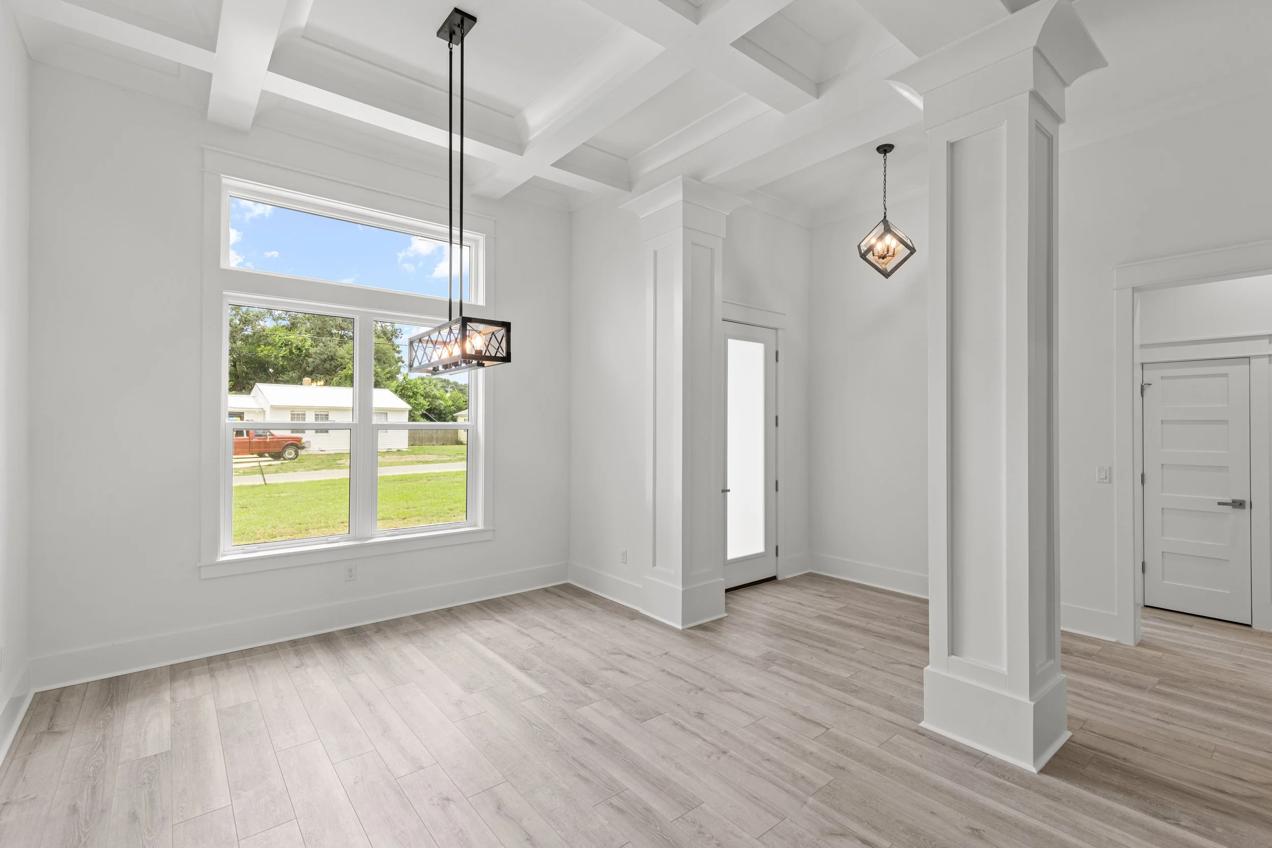 Interior view of a bright, empty living room with large windows, white walls, and wood-look flooring, featuring two modern hanging light fixtures.
