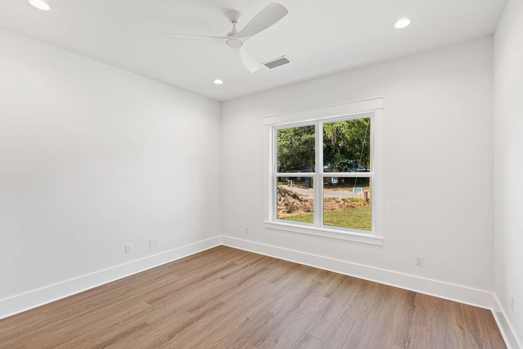Empty room with white walls, wooden flooring, a large window showing an outdoor scene with trees and dirt, a ceiling fan, and soaking up natural light.